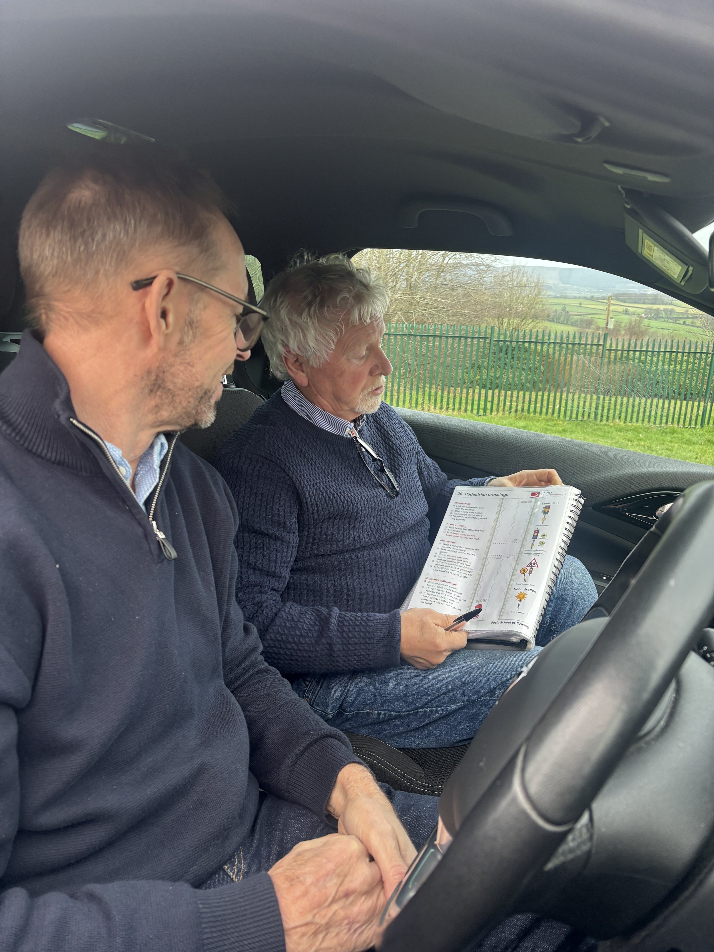Jim Doyle teaching another driving instructor.  The two men are sitting in a car, one holding a pen and a booklet, the other clasping his hands, with a scenic countryside view outside the window.