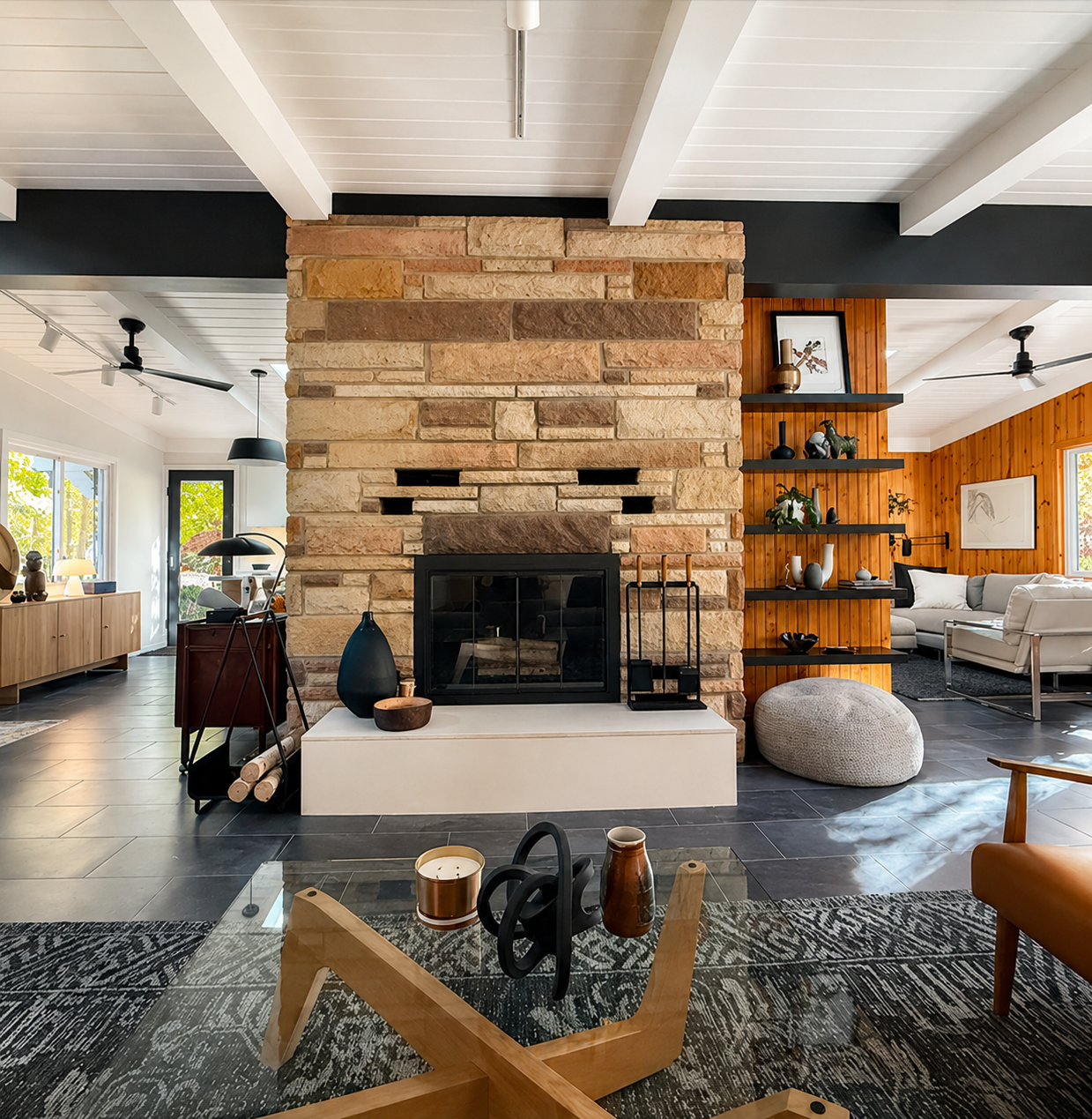 The home's dramatic stone fireplace serves as the centerpiece of this open-concept living space, blending natural textures with clean modern lines. Soaring beamed ceilings, warm wood accents, and thoughtfully curated interiors create a timeless mid-c