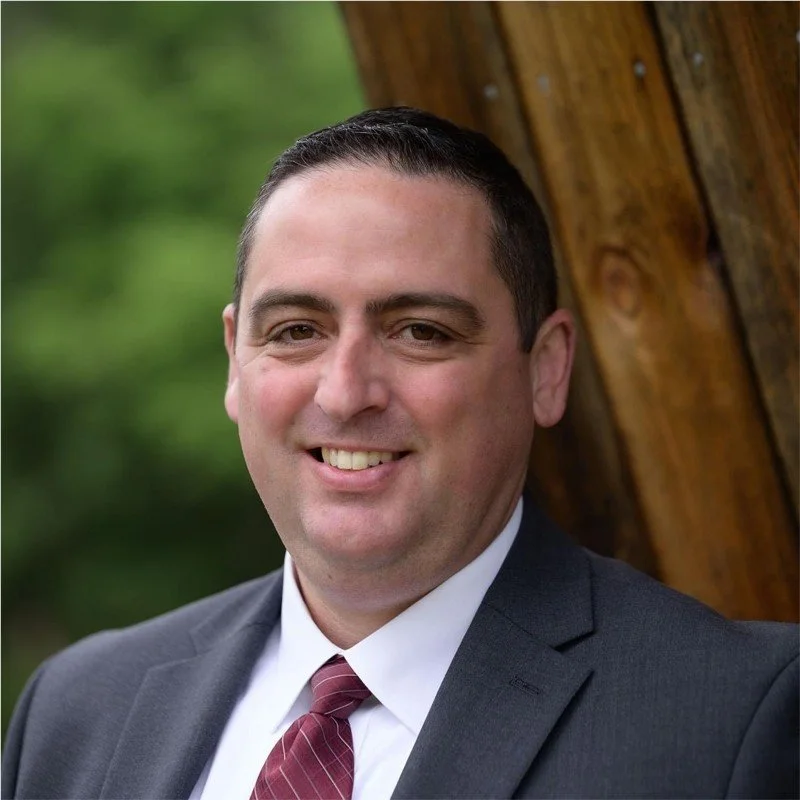 A smiling man in a suit and tie leaning against a wooden structure outdoors with a blurred green background.