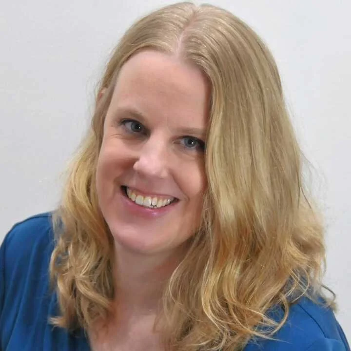 A woman with wavy blonde hair and blue eyes, smiling in front of a plain white background.