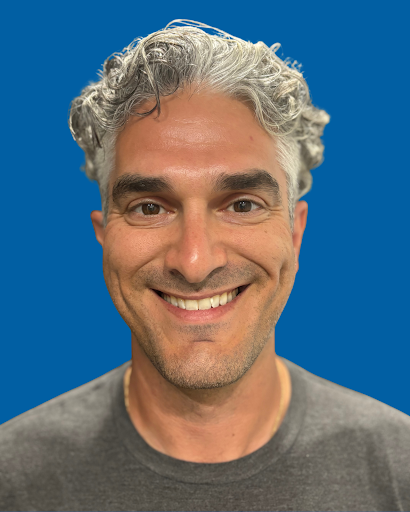 A smiling man with curly gray hair and a beard wearing a dark gray t-shirt, against a solid blue background.