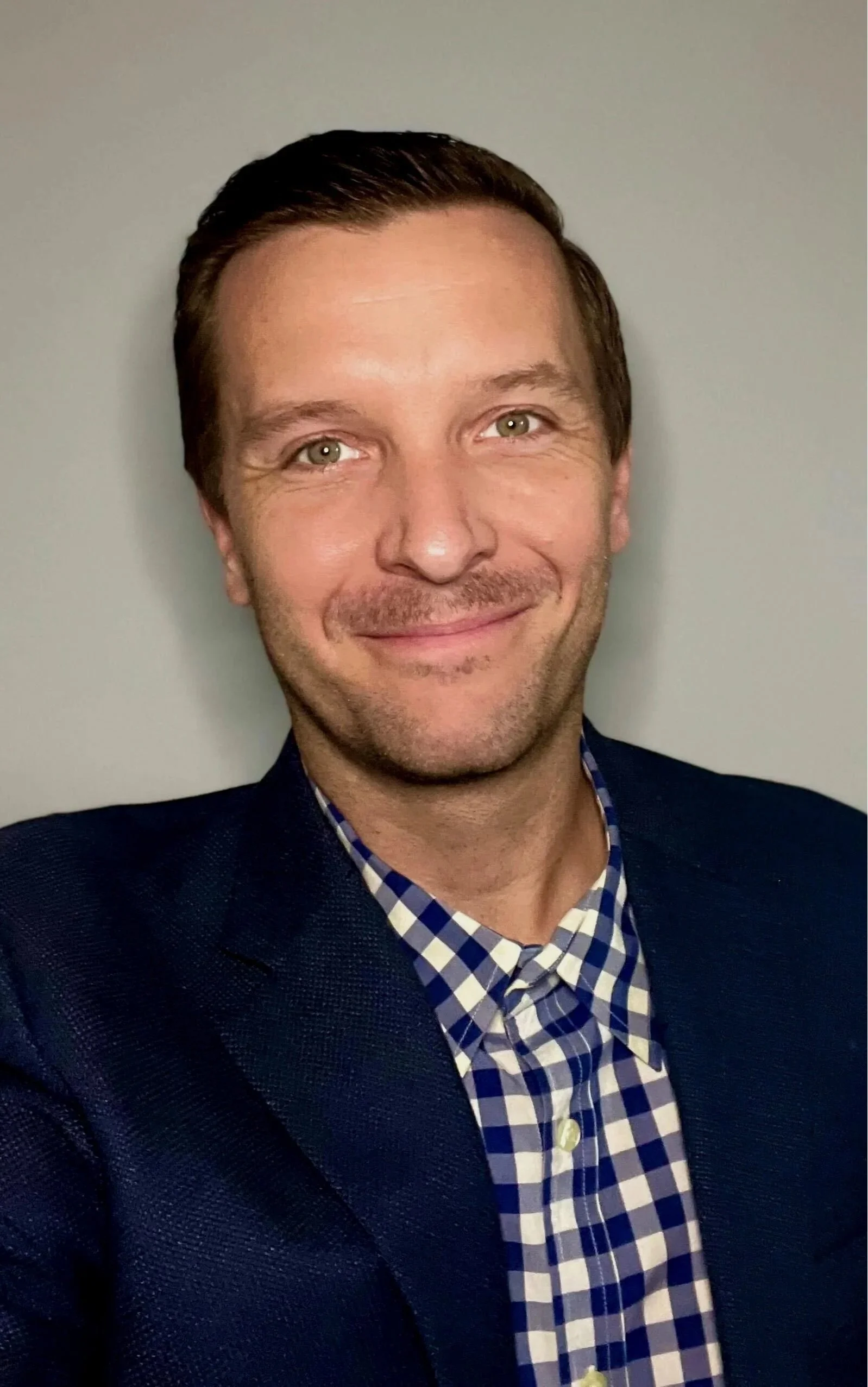 A young man with a beard and short hair wearing a tuxedo with a white bow tie, smiling, standing indoors near a window.