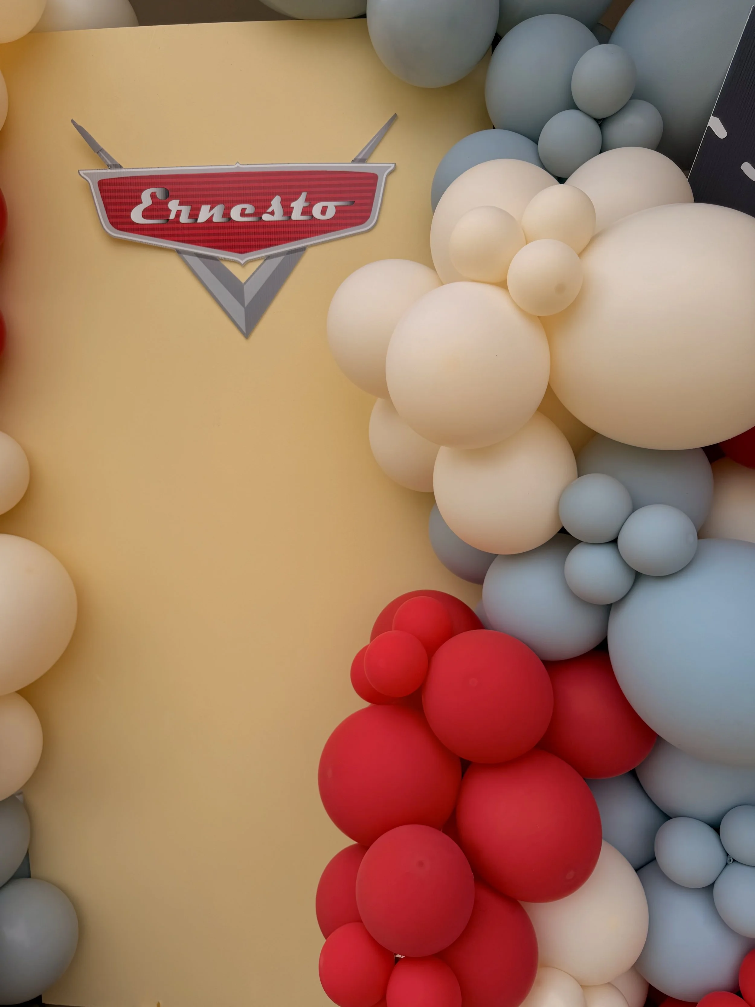 Custom Cars-themed birthday backdrop featuring personalized Ernesto signage inspired by Disney Pixar's Lightning McQueen, framed with organic balloon garlands in red, ivory, and dusty blue. This luxury birthday party installation creates a fun and in