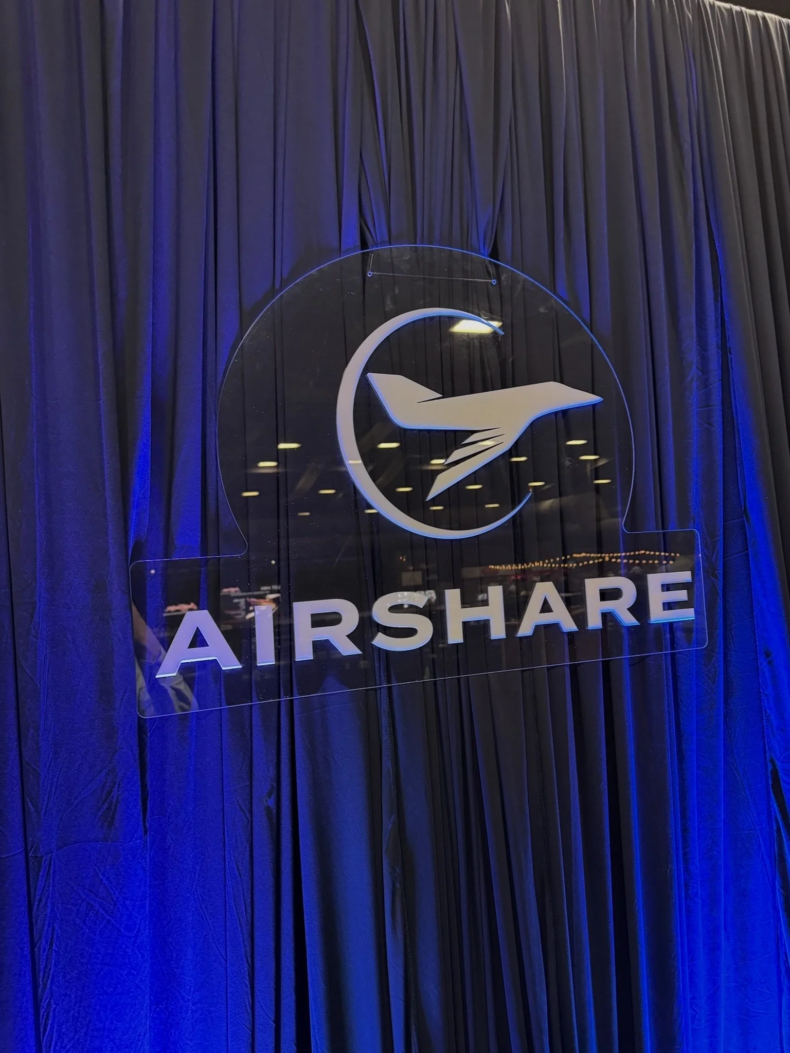 Custom Airshare acrylic logo display featured on luxury navy pipe and drape with blue uplighting for a high-end corporate event experience. Professional event branding and backdrop installation designed for trade shows, executive receptions, VIP loun
