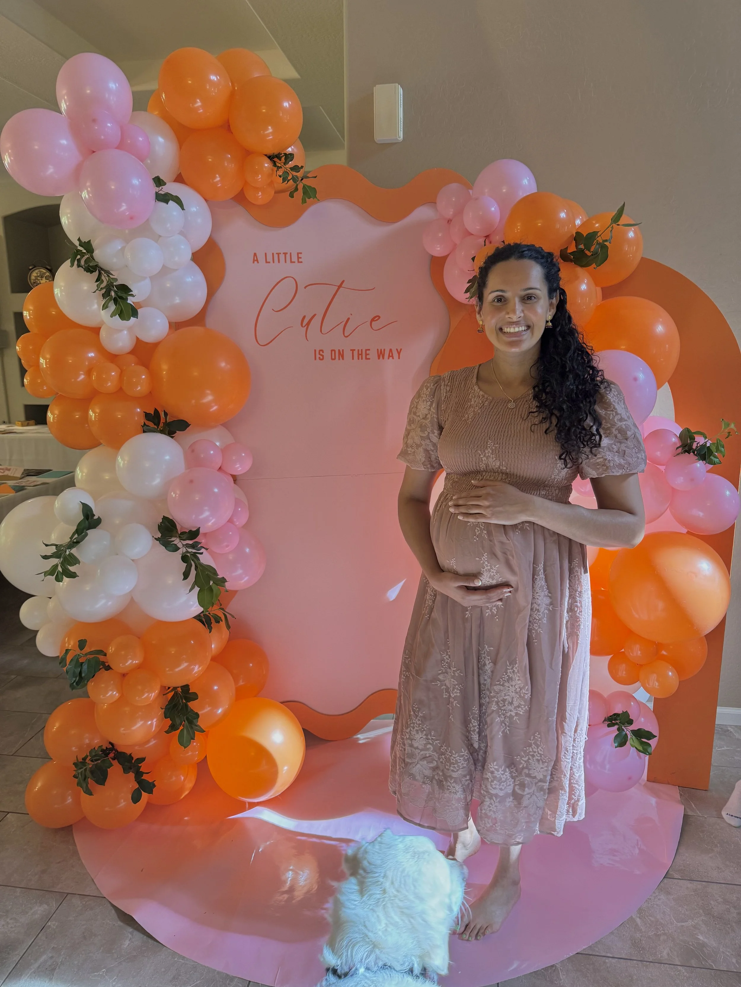Mom-to-be posing in front of a custom "A Little Cutie Is On The Way" baby shower backdrop featuring vibrant orange, blush pink, and white balloon garlands with fresh greenery accents. This modern citrus-inspired baby shower design includes a custom w