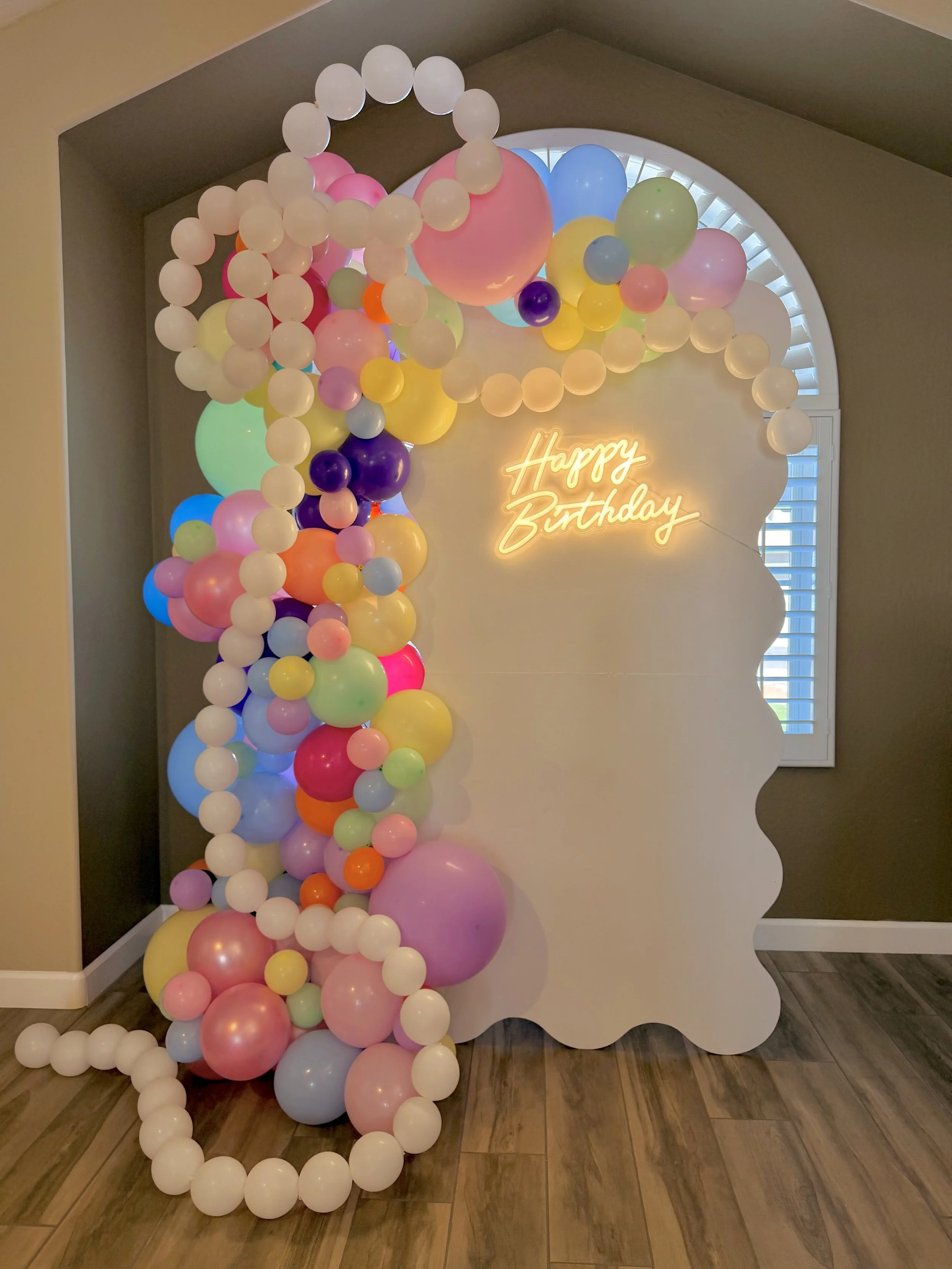 Colorful birthday party backdrop featuring a custom wavy white panel, pastel rainbow balloon garland, glowing "Happy Birthday" neon sign, and whimsical balloon arch details. Designed with playful pastel shades of pink, blue, yellow, purple, green, an
