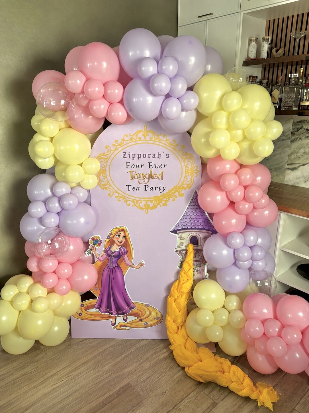 Custom Rapunzel-inspired princess birthday backdrop featuring pastel pink, lavender, and buttercream balloon garlands, a personalized "Zipporah's Four Ever Tangled Tea Party" sign, whimsical castle details, and a cascading golden braid. This enchanti