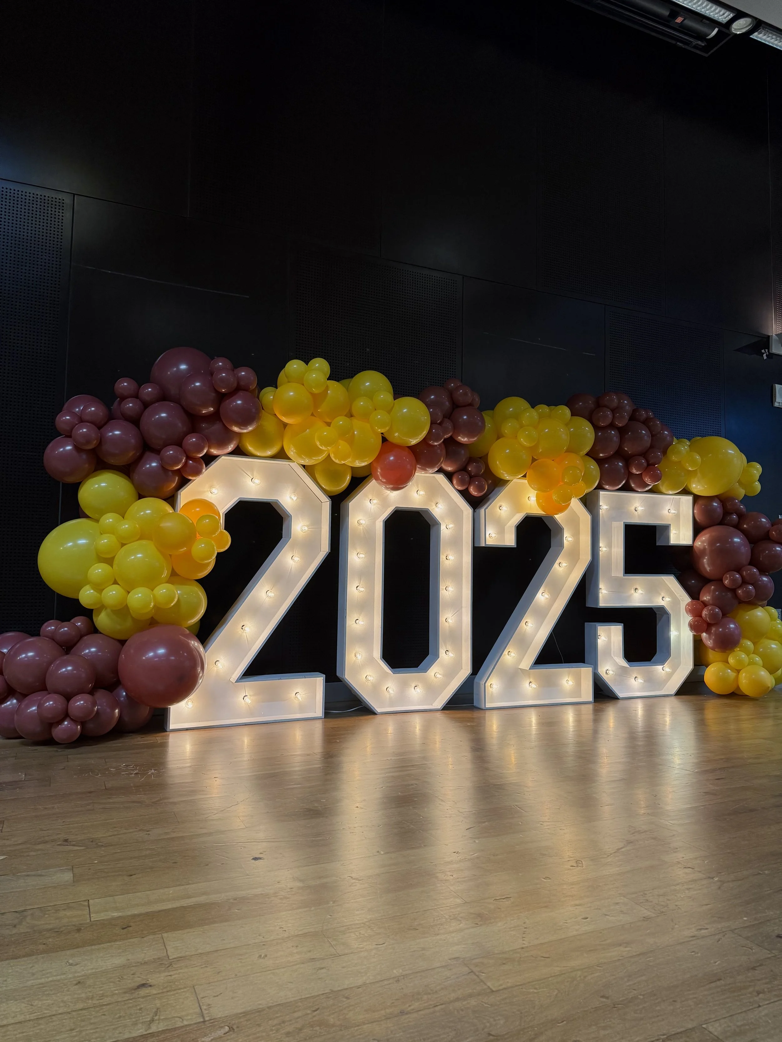 Custom graduation event backdrop featuring illuminated 2025 marquee numbers and organic maroon and gold balloon garlands inspired by Arizona State University colors. Designed for commencement ceremonies, graduation celebrations, senior recognition ev