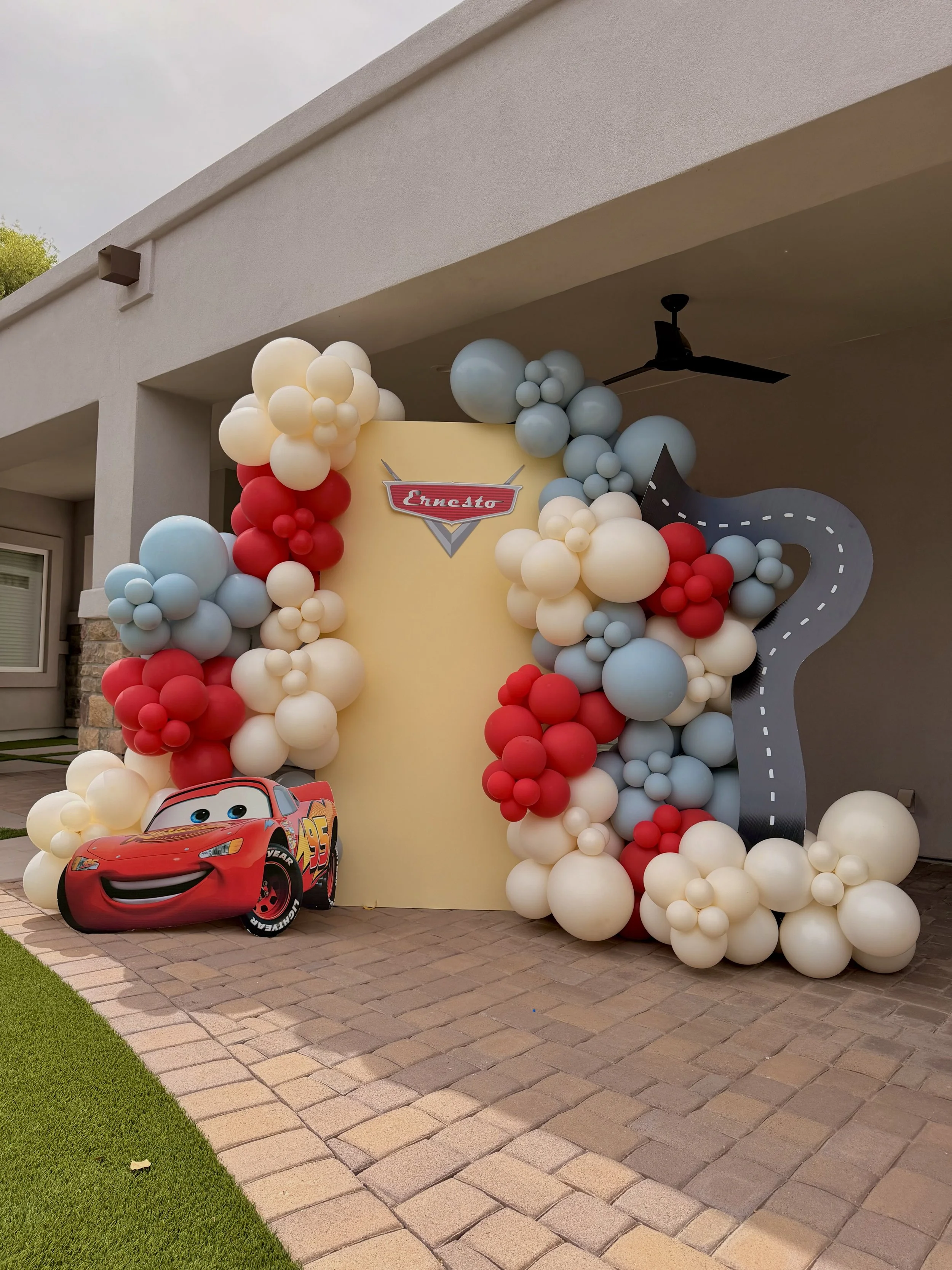Custom Disney Pixar Cars birthday party backdrop featuring Lightning McQueen, race track décor, personalized event signage, and organic balloon garlands in red, blue, ivory, and white. Designed for Ernesto's birthday celebration, this interactive pho