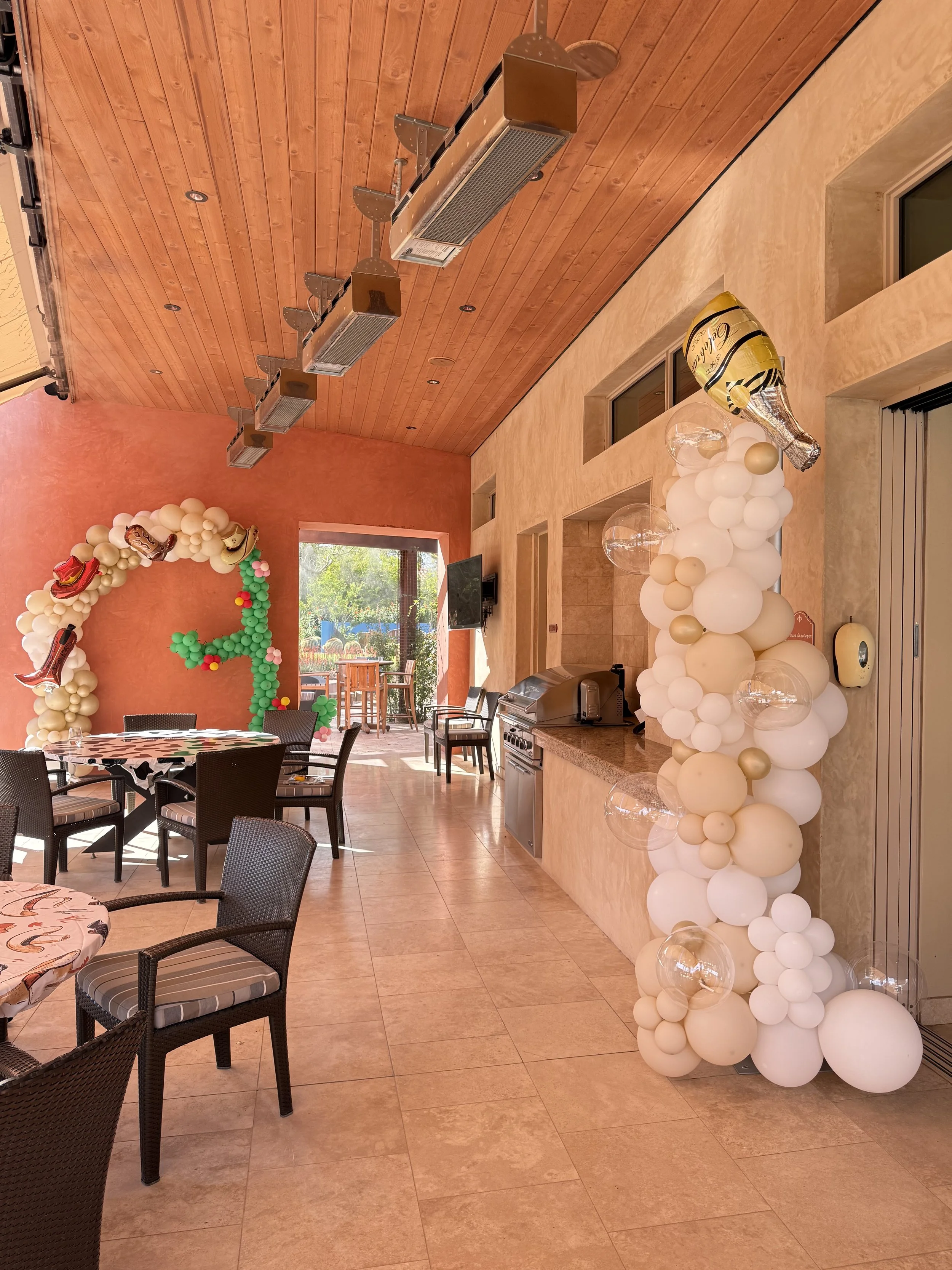 Custom western-themed birthday party décor featuring an organic white and champagne balloon column with bubble balloons, champagne bottle accents, and a Wild West-inspired balloon backdrop with cowboy boots, hats, and cactus details. Designed for lux