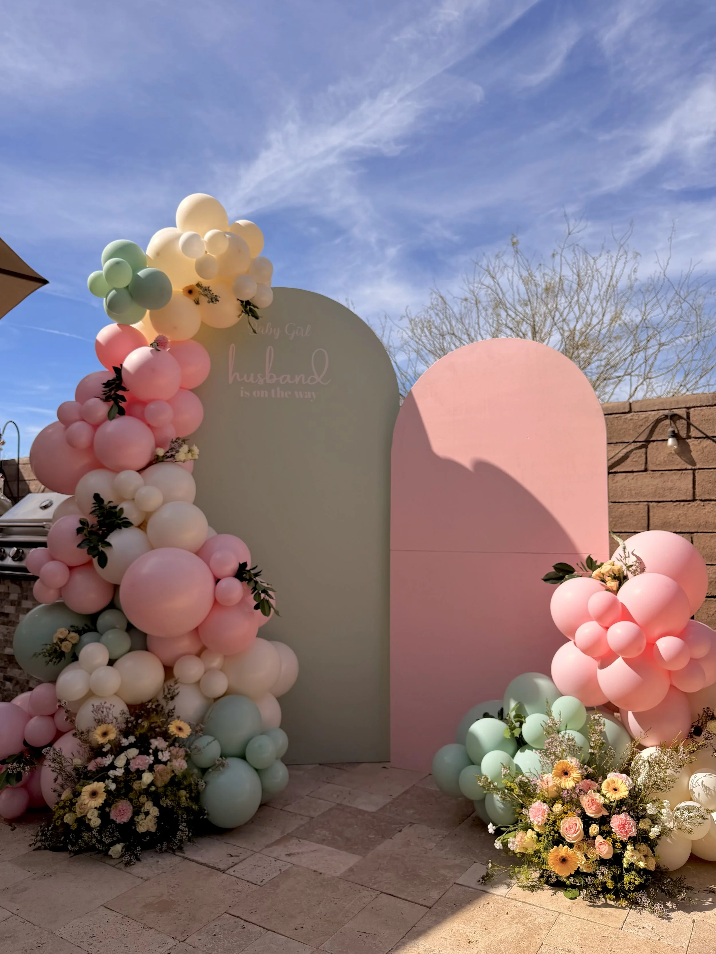 Luxury baby shower backdrop featuring custom arched panels, pastel pink, mint green, ivory, and white balloon garlands, and garden-inspired floral arrangements. This elegant baby shower installation combines organic balloons with fresh florals to cre