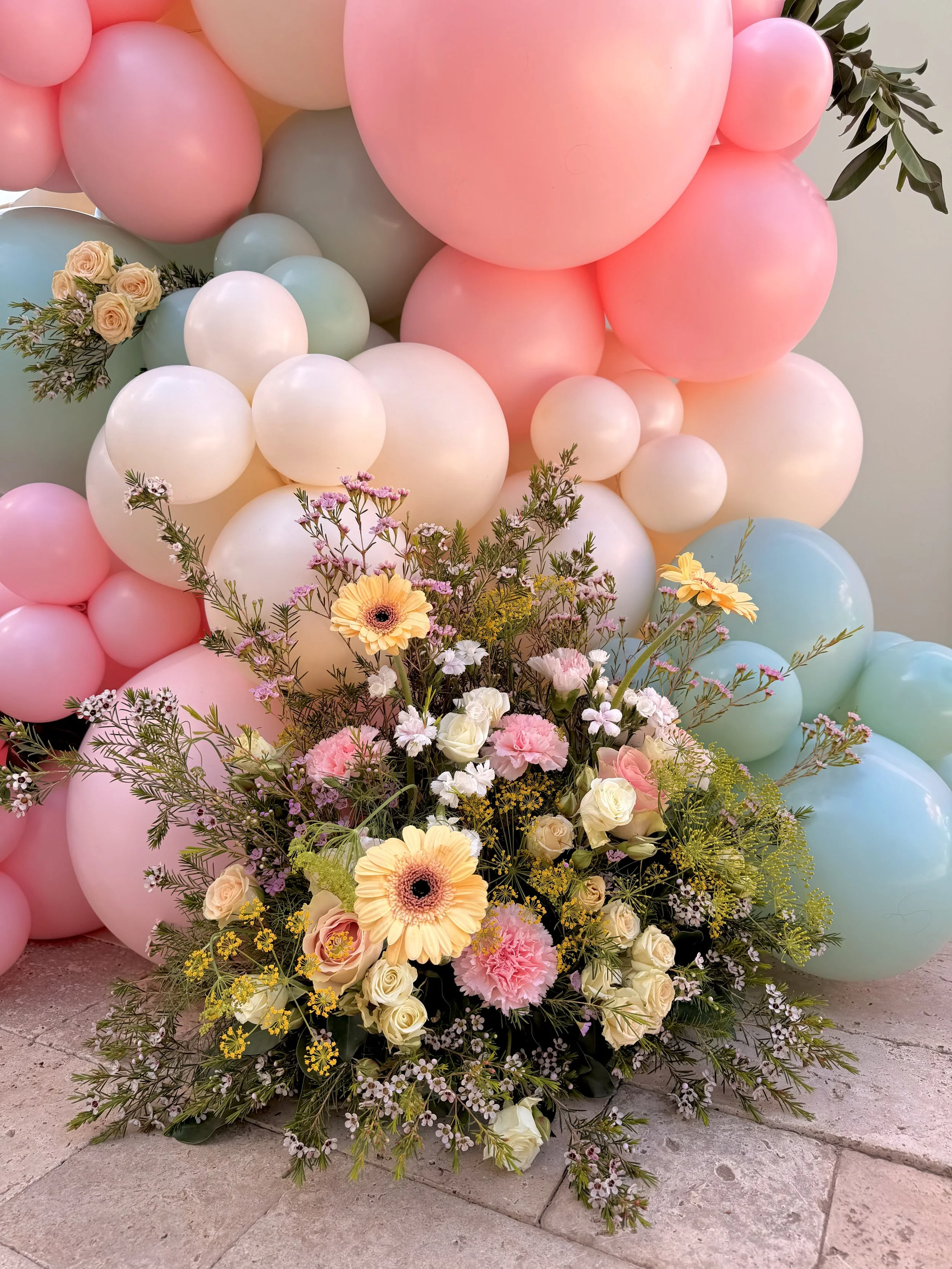 Whimsical garden-inspired floral arrangement paired with a pastel balloon garland featuring blush pink, ivory, mint, and baby blue balloons. Designed with fresh roses, gerbera daisies, carnations, wax flower, and seasonal blooms, this luxury event in