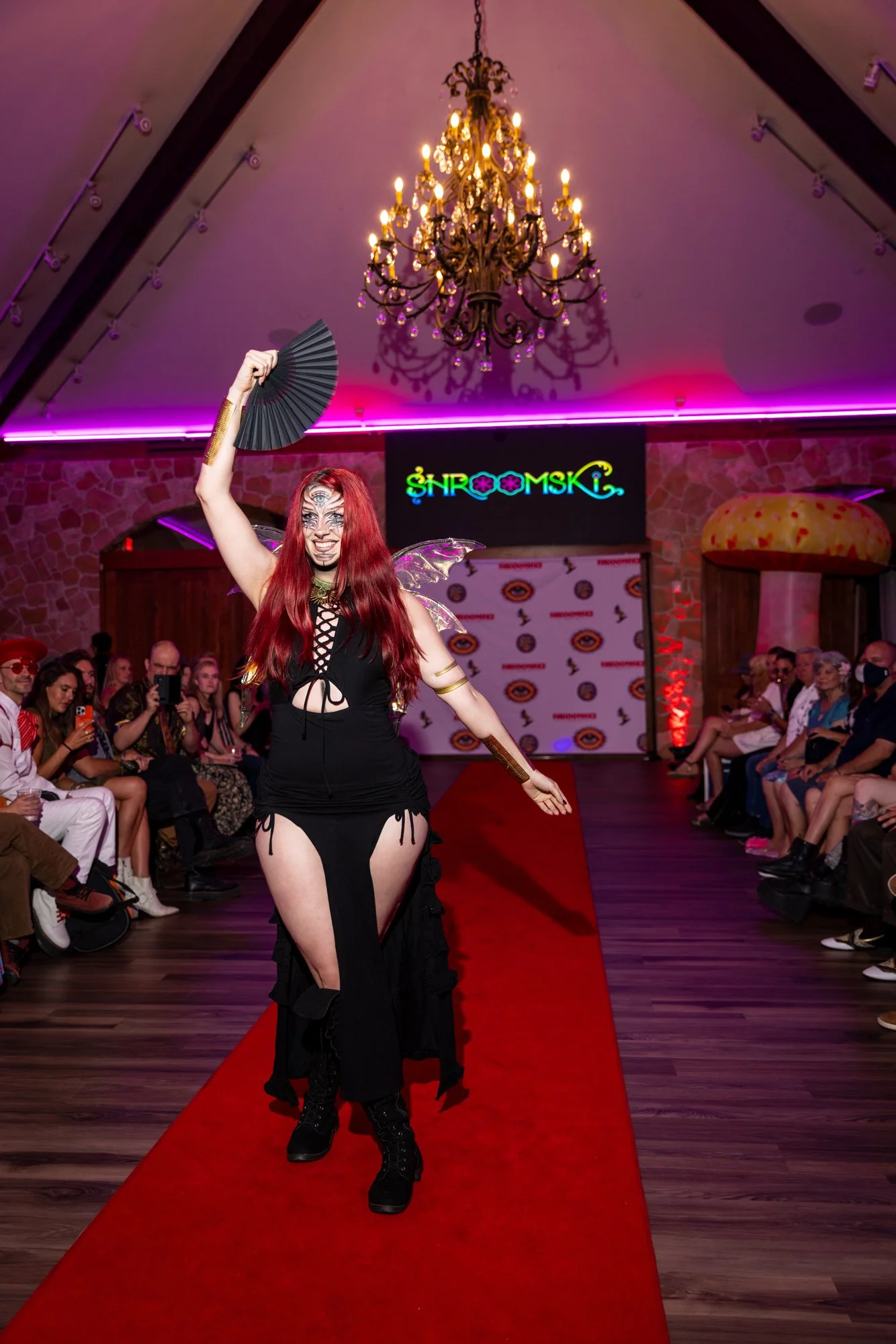 A performer with red hair dressed in black, holding a fan in one hand, walking down a red carpet runway during a lively event with an audience. The setting includes a chandelier hanging from the ceiling, colorful lighting, and a backdrop with the wor