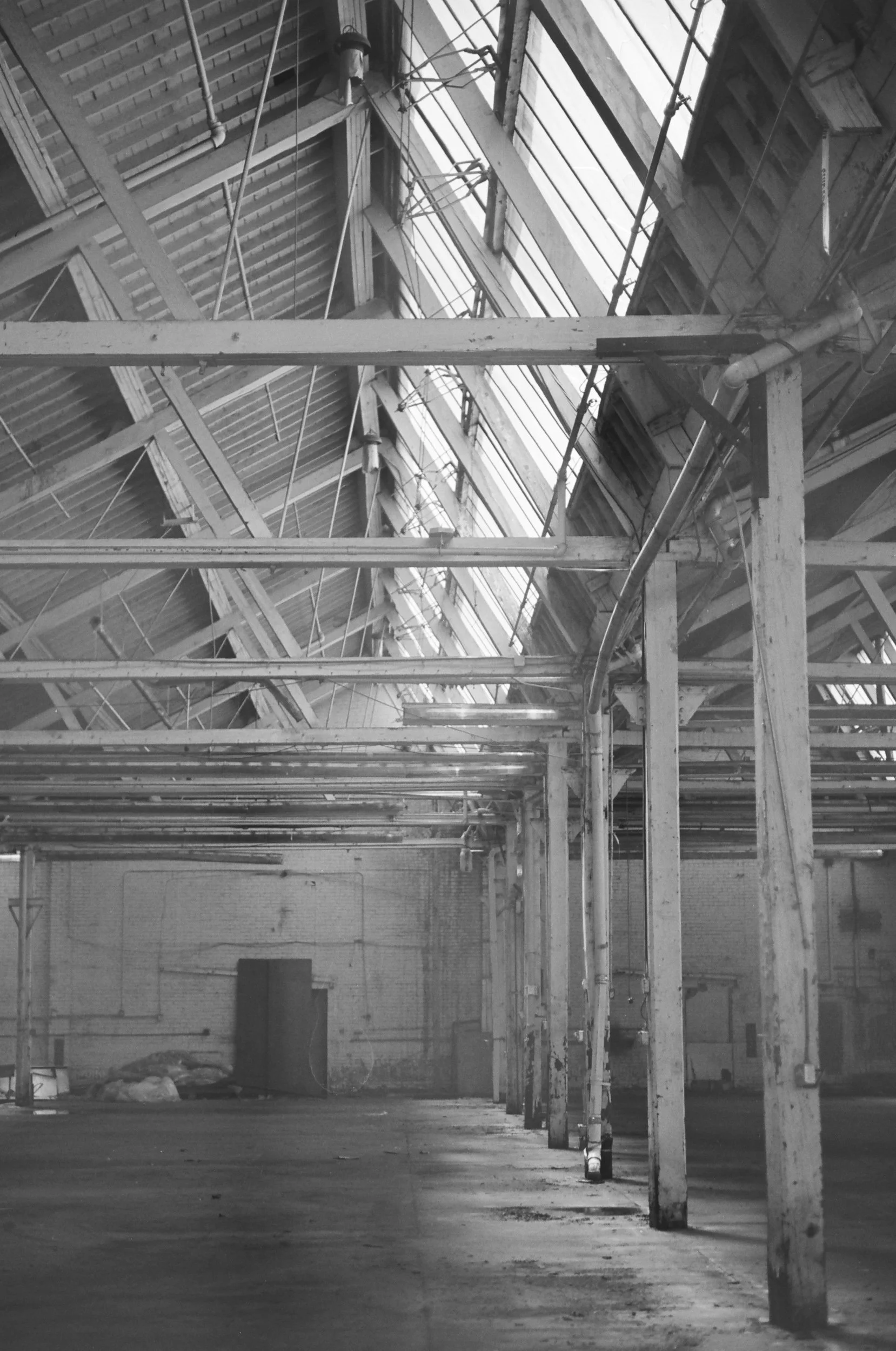 Empty industrial warehouse with exposed wooden beams and ceiling, lit by large windows, with scattered equipment and structural elements.