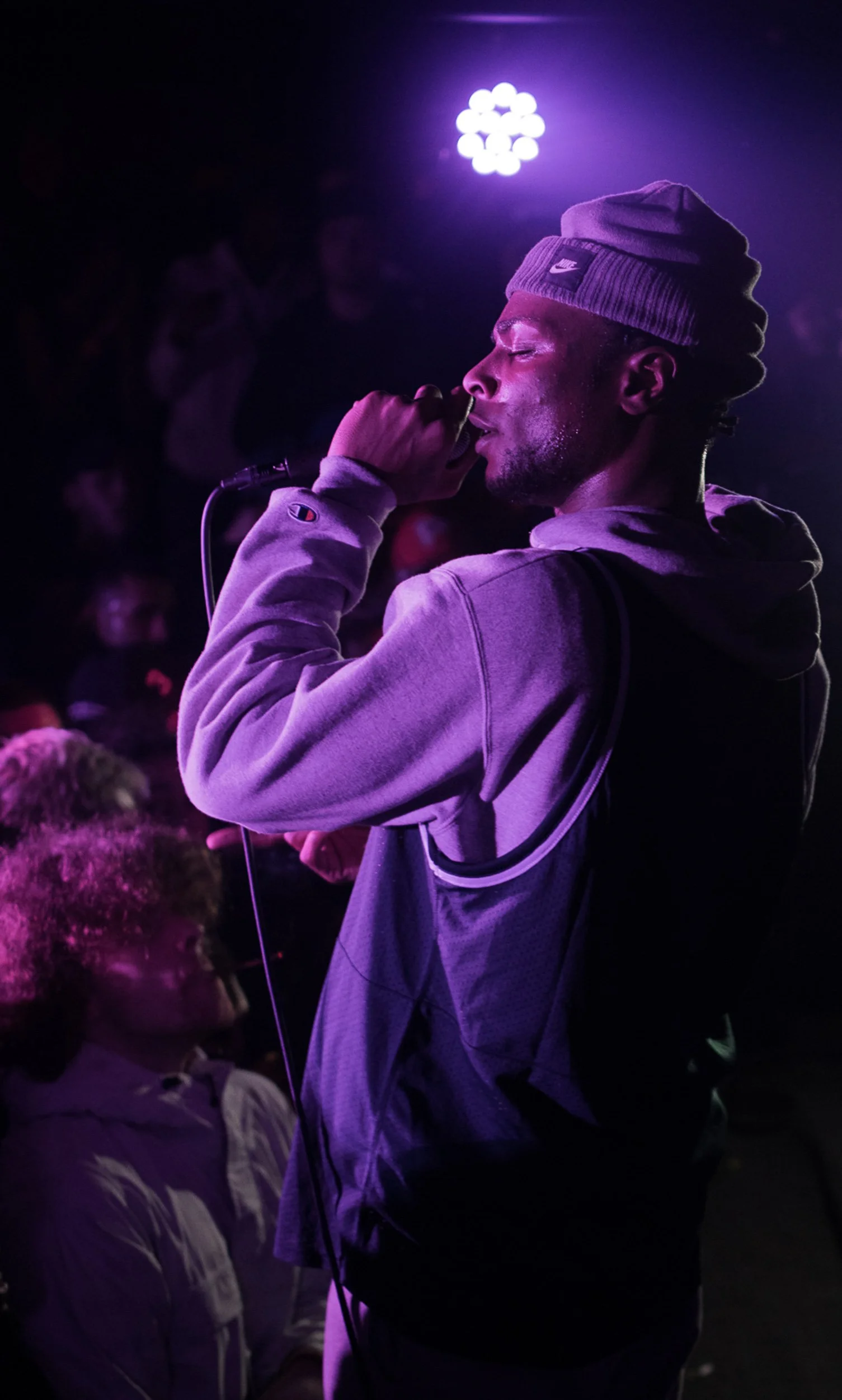 A man with closed eyes singing into a microphone at a dark, purple-lit concert or event, wearing a gray beanie and hoodie, with audience members in the background.