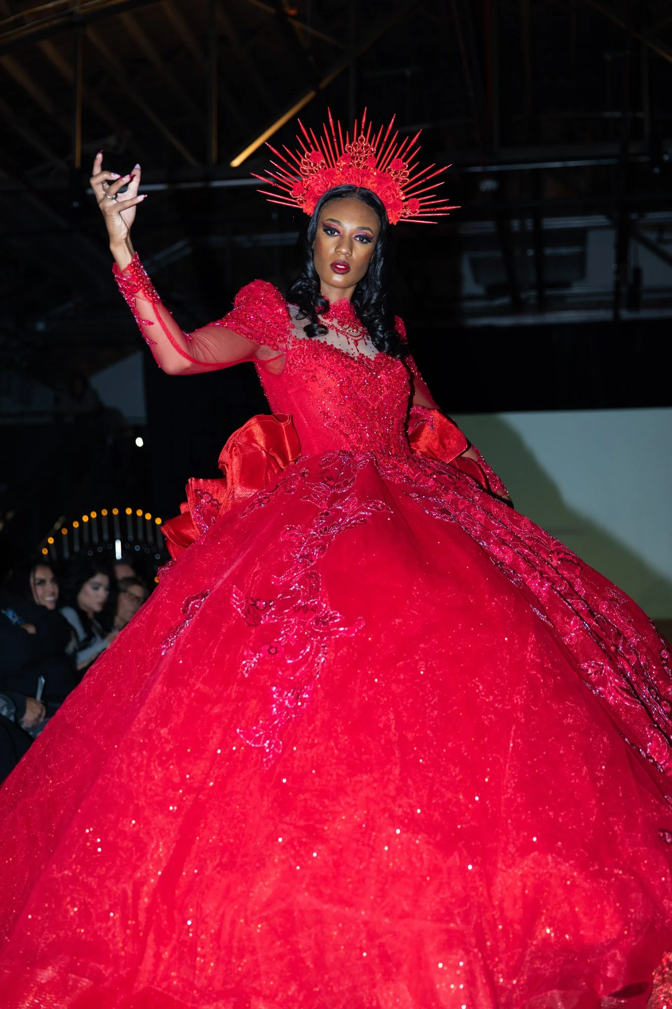 A woman wearing an elaborate, sparkling red gown and a matching red headpiece with spikes and roses, standing on a runway during a fashion show.