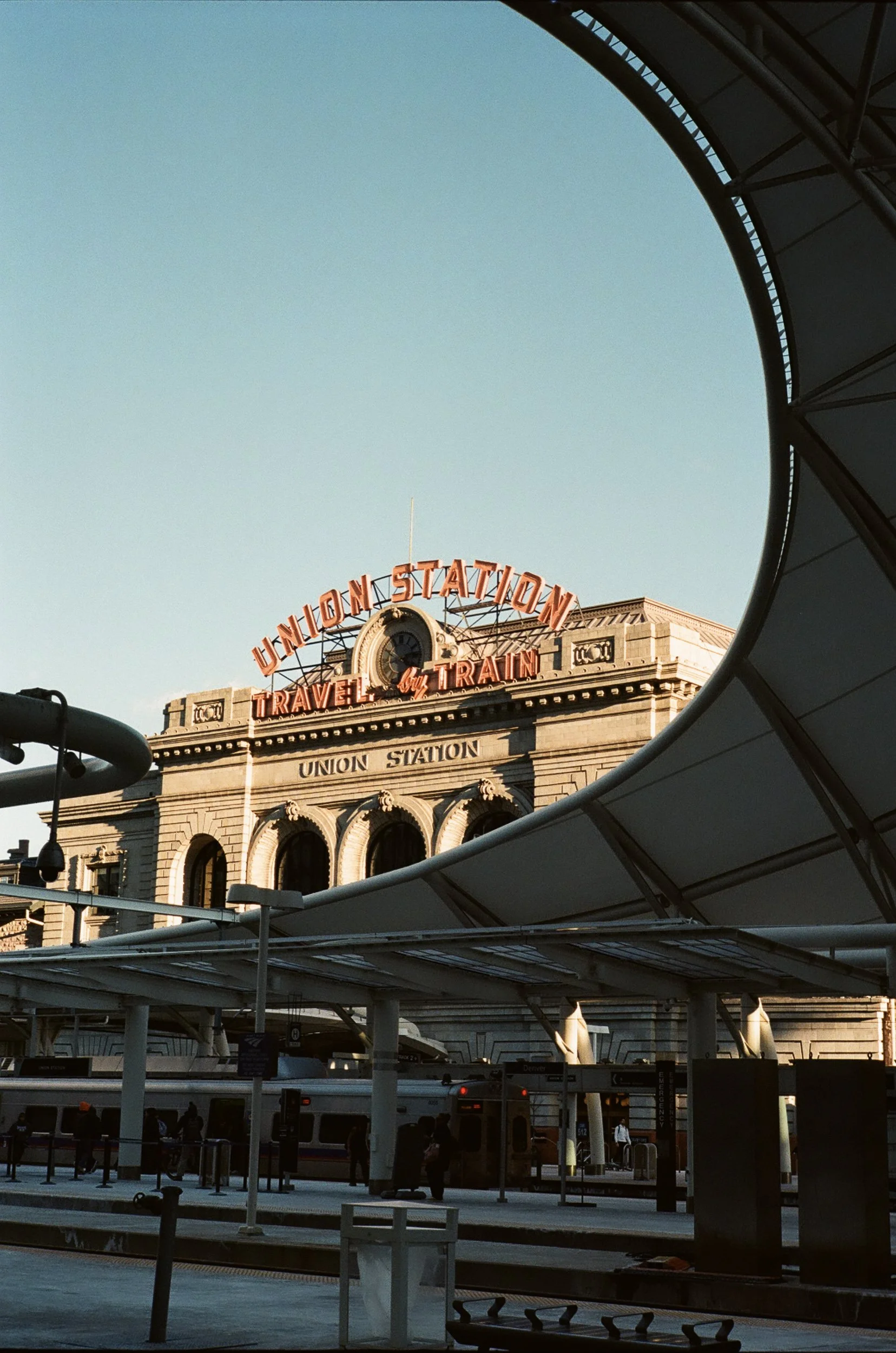 Union Station train station with an outdoor area, visible clock, people walking, a train at the platform, and a large sign reading 'Union Station' with the slogan 'Travel by Train' underneath.