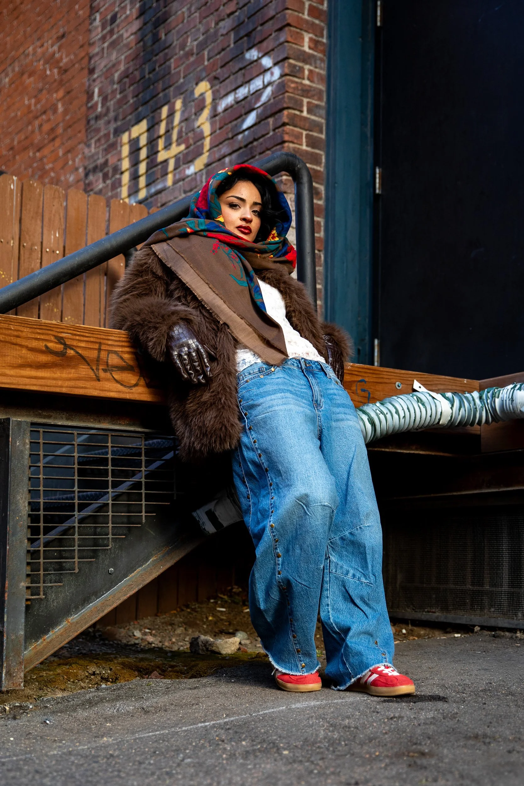 Young woman leaning on a railing in an urban alleyway with brick walls, wearing a colorful headscarf, fur-lined coat, jeans, and red sneakers.