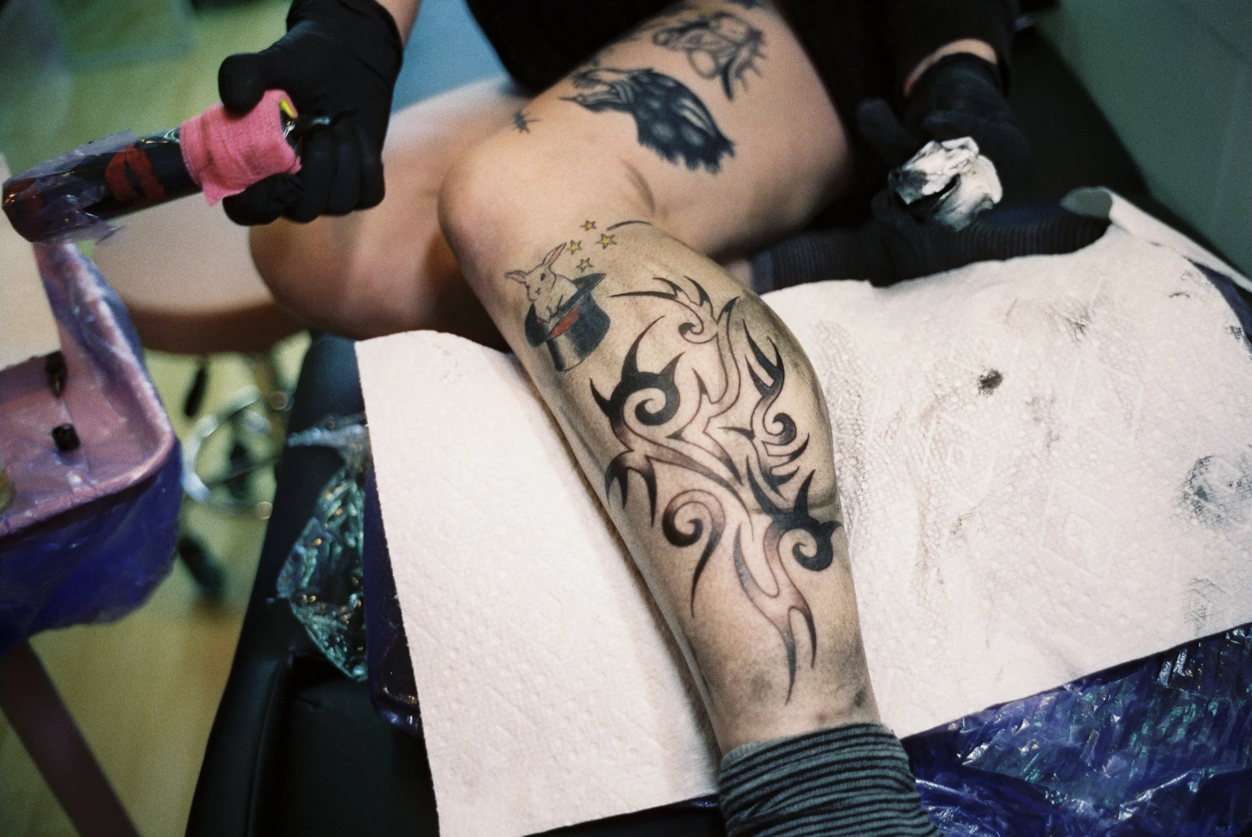 A person getting a tattoo on their leg, with the tattoo featuring a rabbit in a magician's hat and stars. The person is seated on a tattoo chair, wearing black gloves and striped socks, with a towel covering parts of their body. The tattoo artist is 