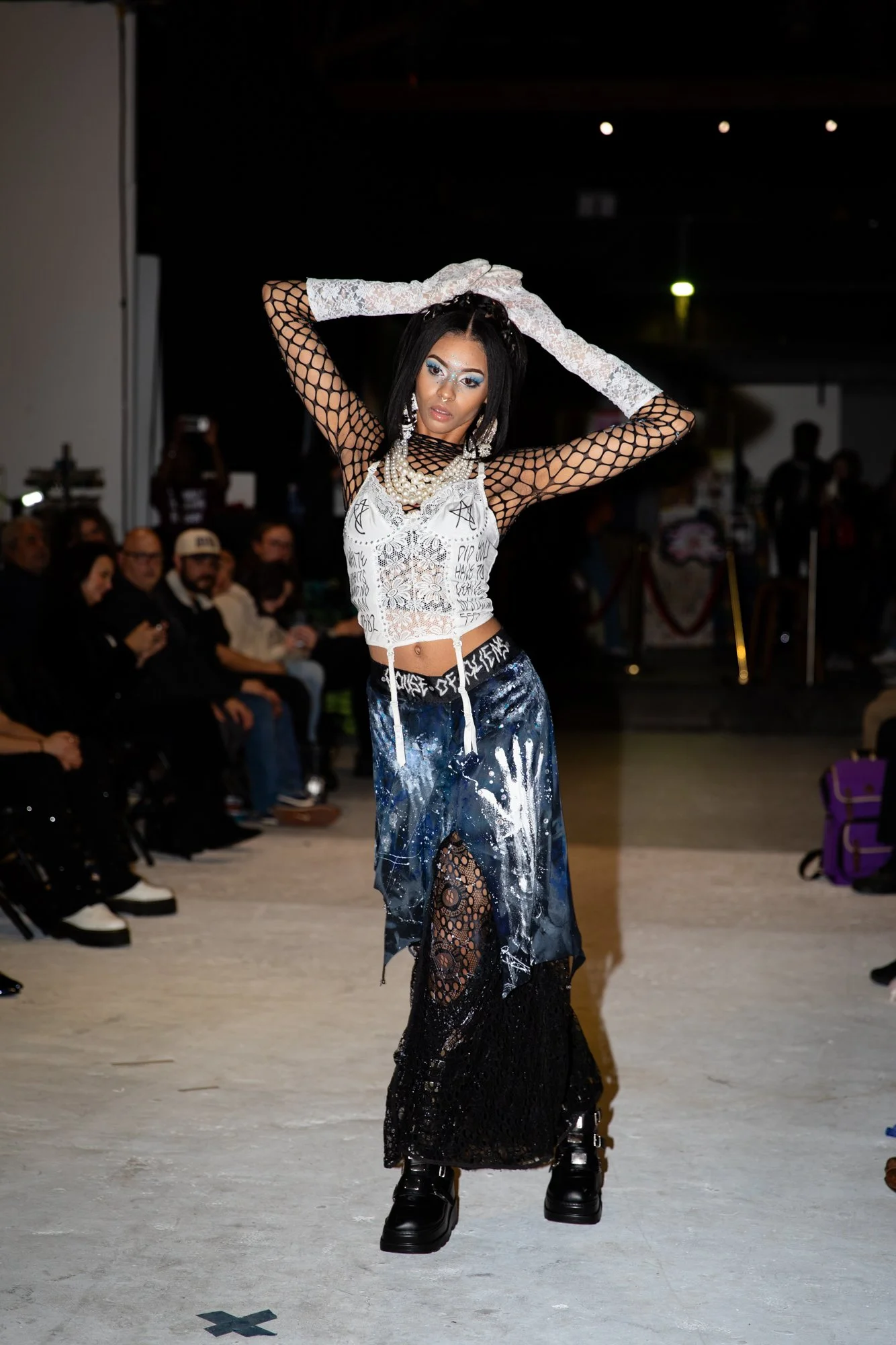 A fashion model walking on a runway during a fashion show, wearing a black mesh top with lace gloves, layered pearl jewelry, a white lace crop top with graffiti-like writings, galaxy-themed pants, black lace and platform boots, with audience members 