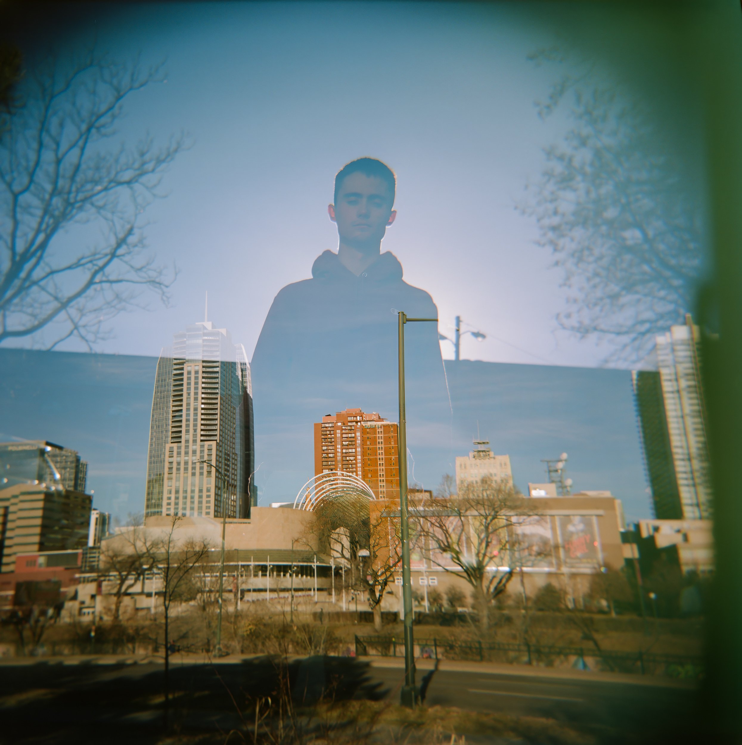 Reflection of a man with short hair wearing a hoodie in a body of water with trees, a cityscape, and buildings in the background.