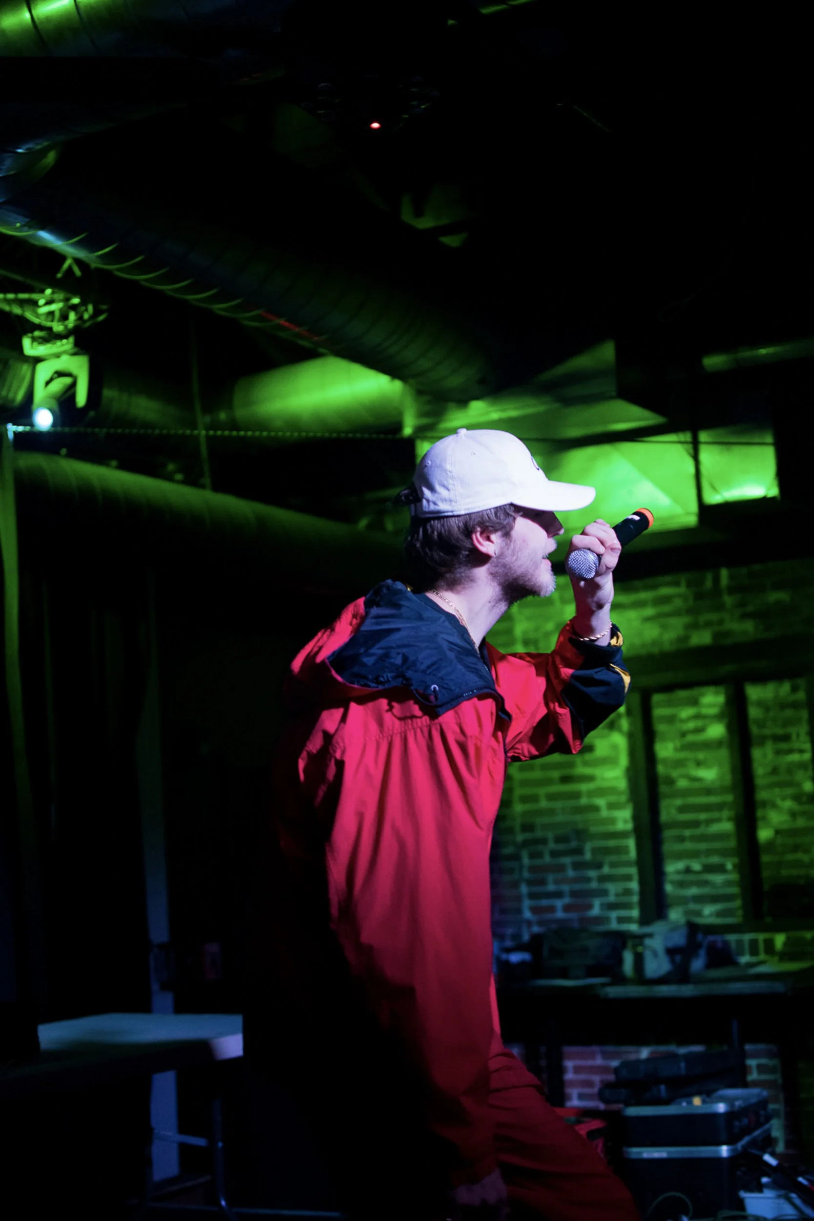 A person wearing a white cap and red jacket holding a microphone, performing on stage with green and blue lighting, and a brick wall in the background.