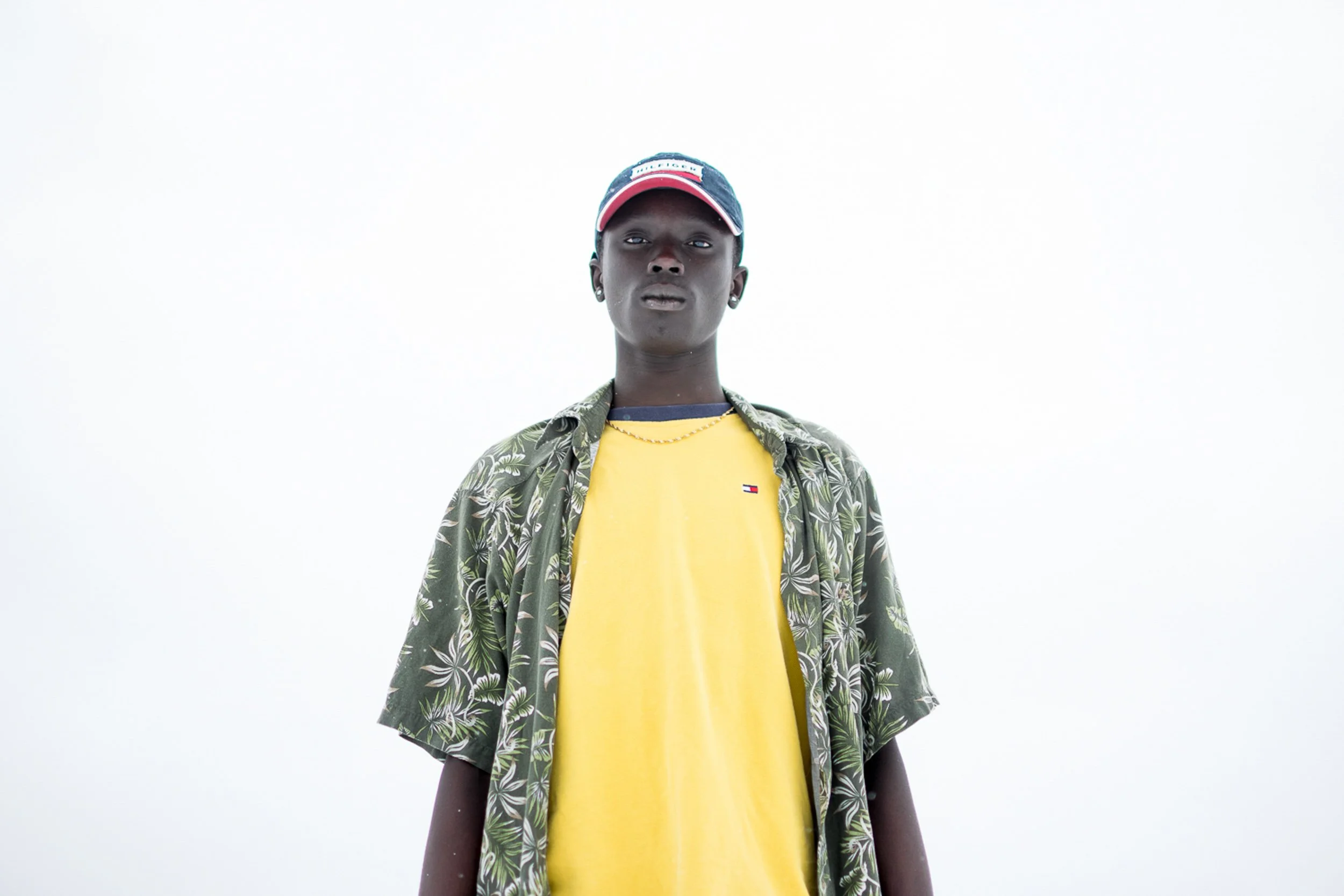 A person with dark skin wearing a yellow T-shirt, a patterned shirt, a baseball cap, and earrings stands against a plain white background.
