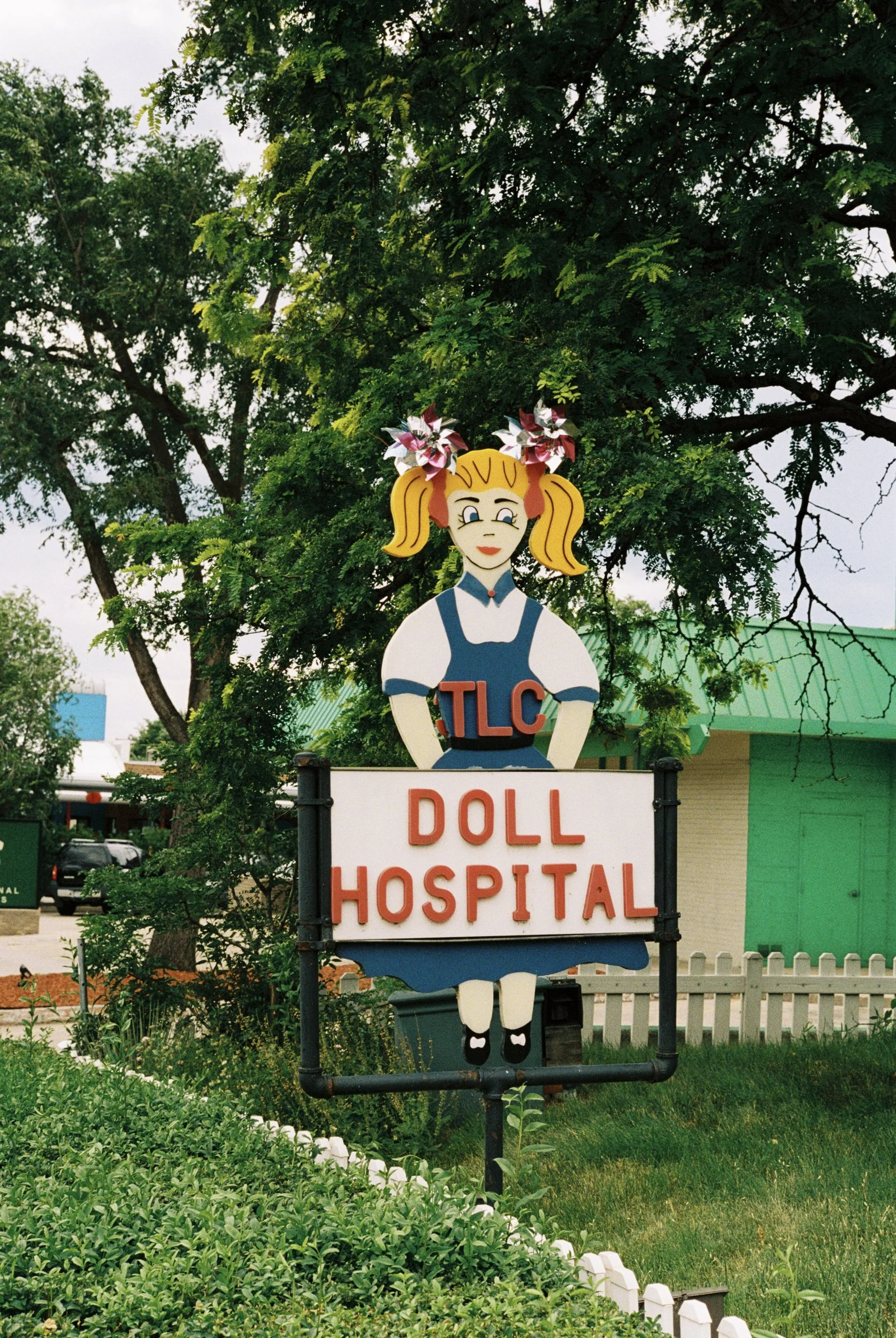 Sign for a doll hospital with a cartoon doll figure at the top, surrounded by trees and greenery.