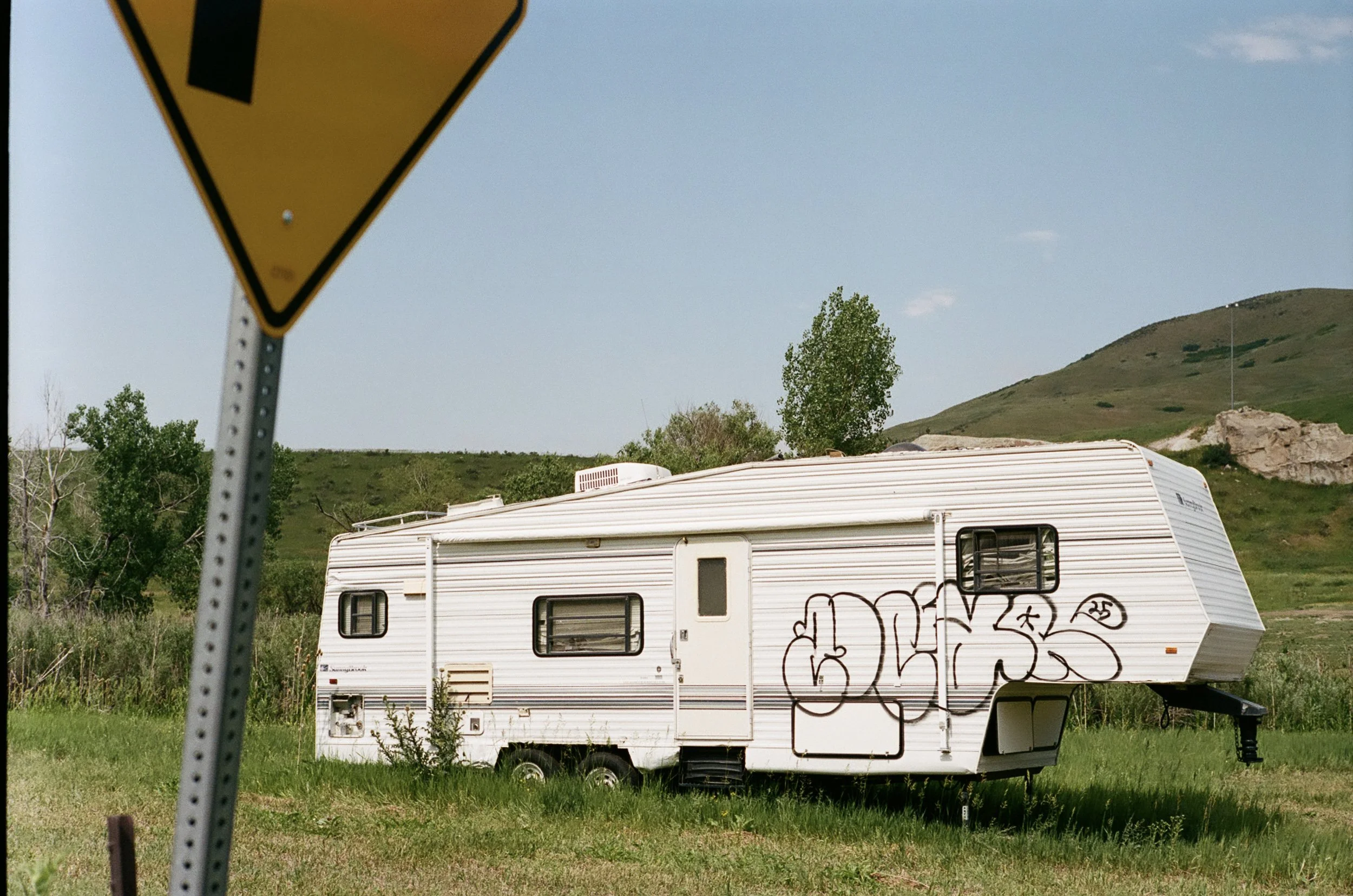 A white camper trailer with graffiti on the side, situated on a grassy area with sparse bushes, green hills, and a blue sky in the background. A yellow traffic sign with a black exclamation mark is partially visible in the foreground.