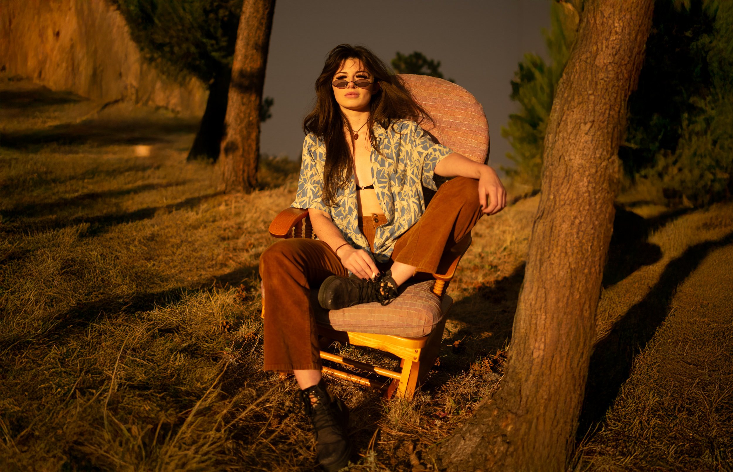 A young woman with dark hair wearing sunglasses, a patterned shirt, and brown pants sitting on a chair outdoors next to trees at dusk.