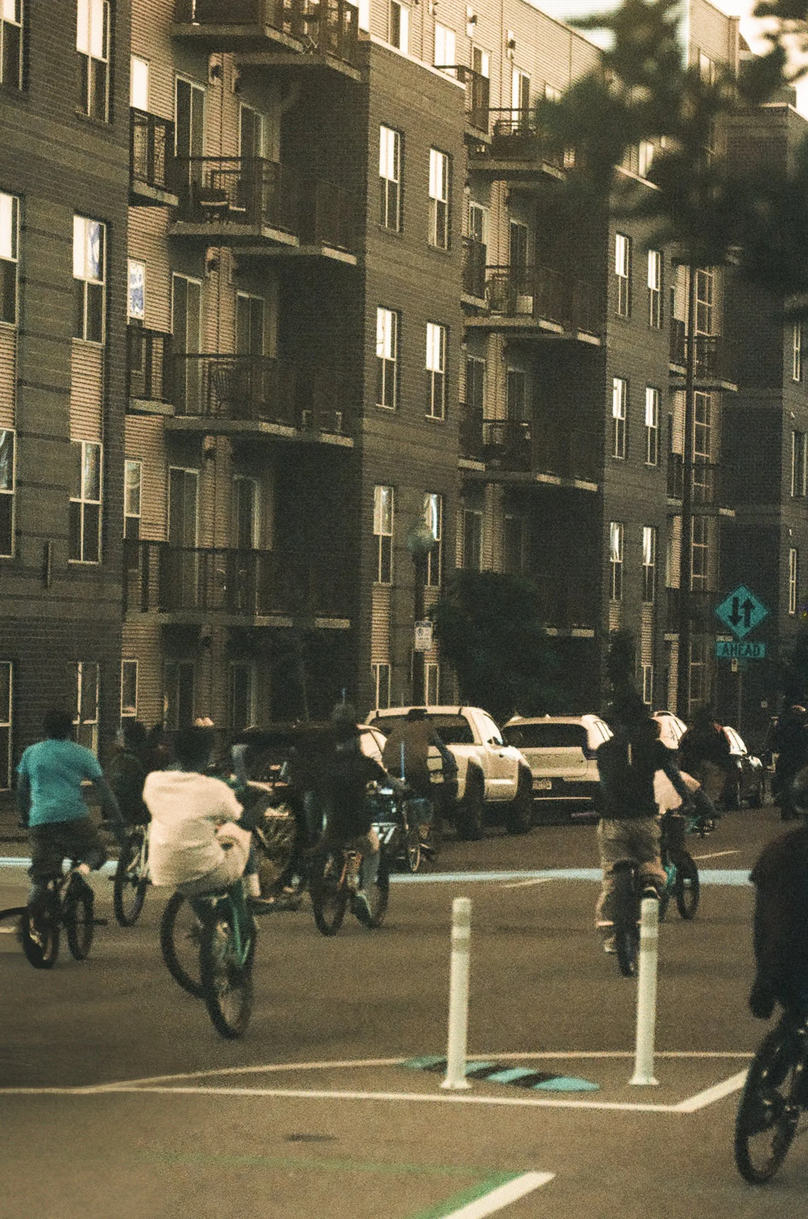 Group of people riding bicycles and skateboards in a parking lot with parked cars and an apartment building in the background.