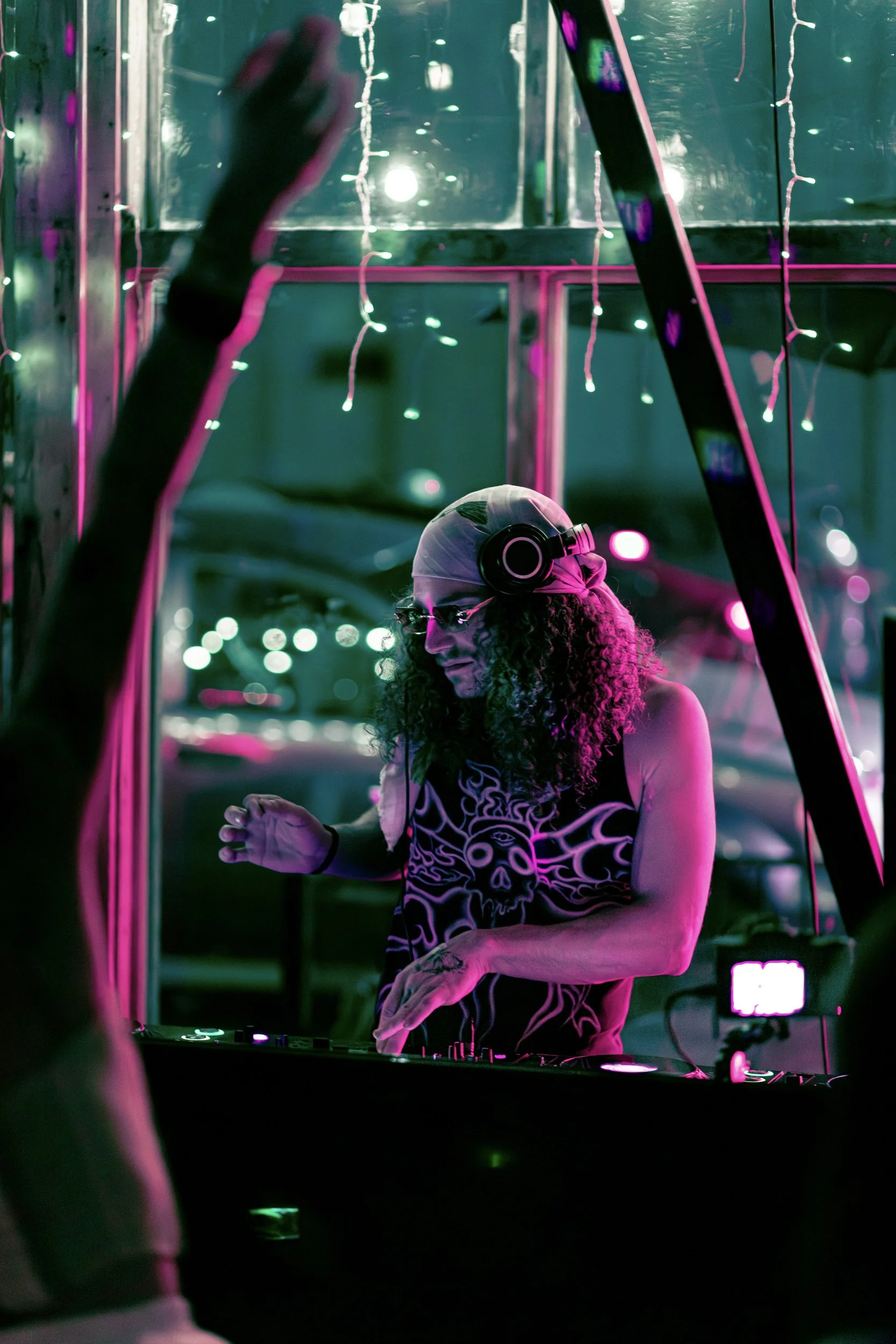 A person with curly hair wearing headphones and a skull-themed shirt, DJing at a neon-lit event with pink and green lighting, and fairy lights in the background.