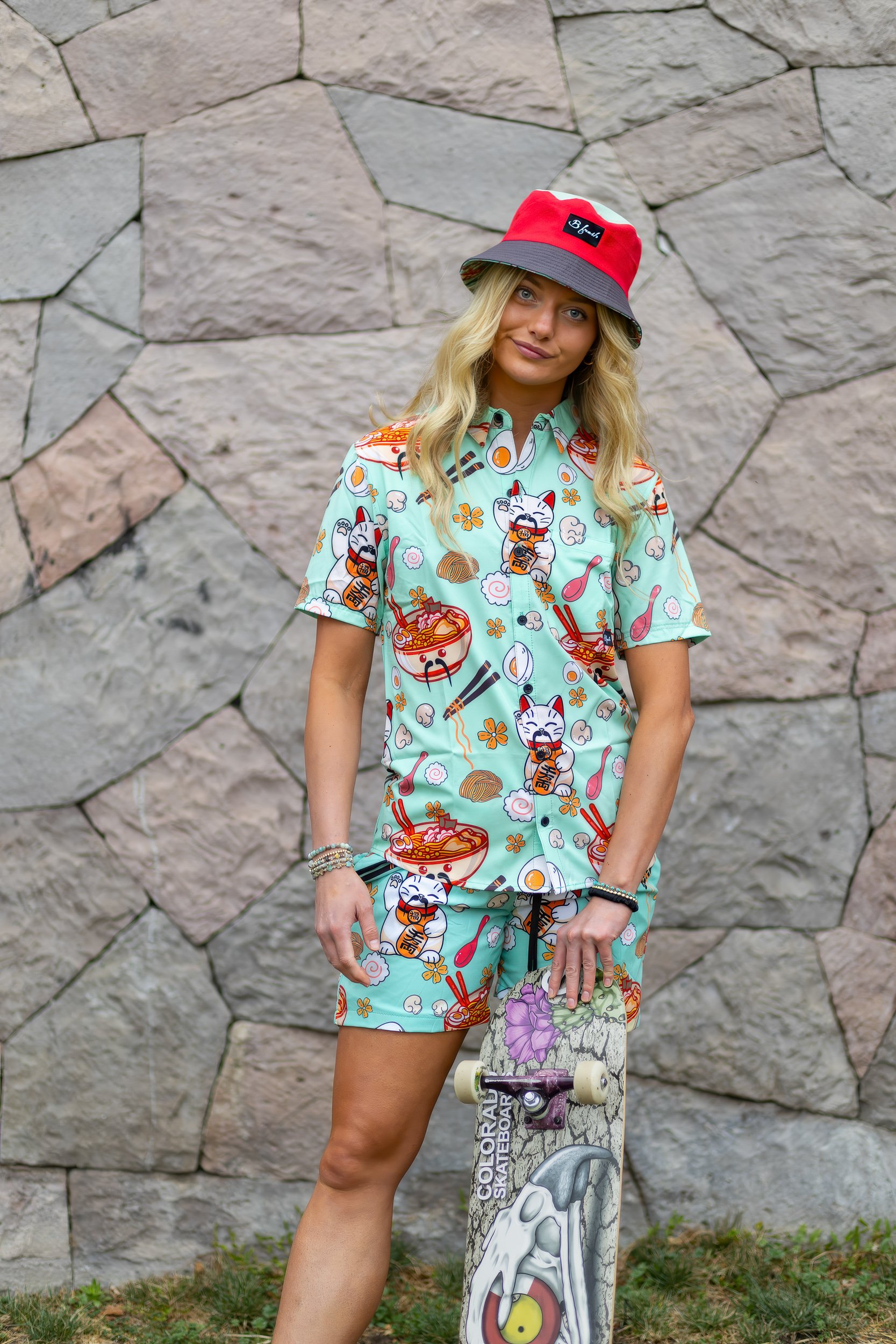 A young woman with long blonde hair wearing a colorful patterned shirt and shorts, a red and gray bucket hat, and bracelets, standing outdoors against a stone wall, holding a skateboard.