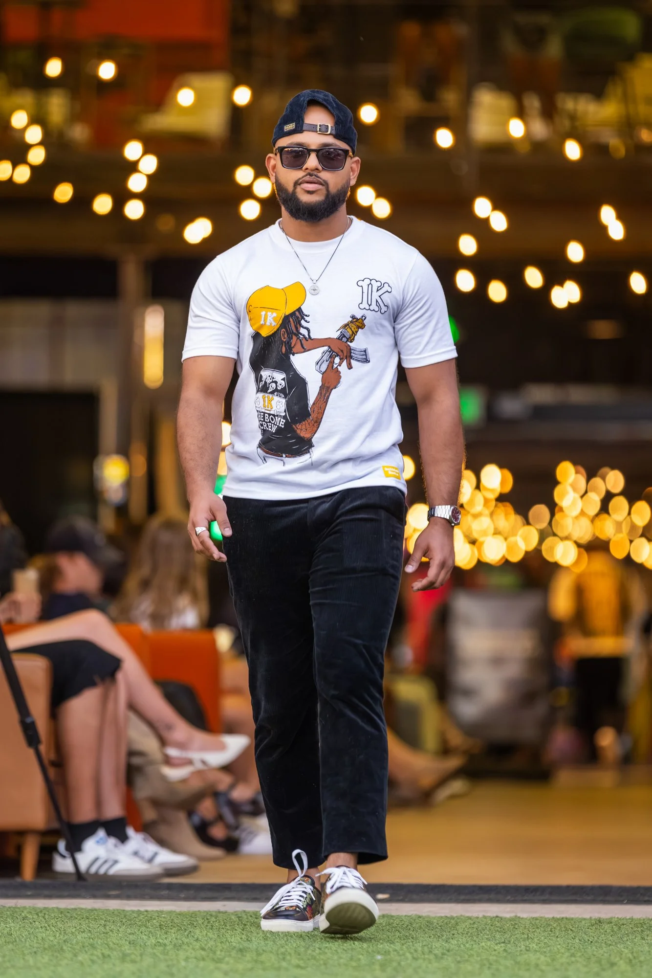 A man with a beard wearing sunglasses, a backward baseball cap, a white graphic t-shirt, black pants, and sneakers walking on a green surface in a brightly lit indoor space with blurred background lights and seated people.