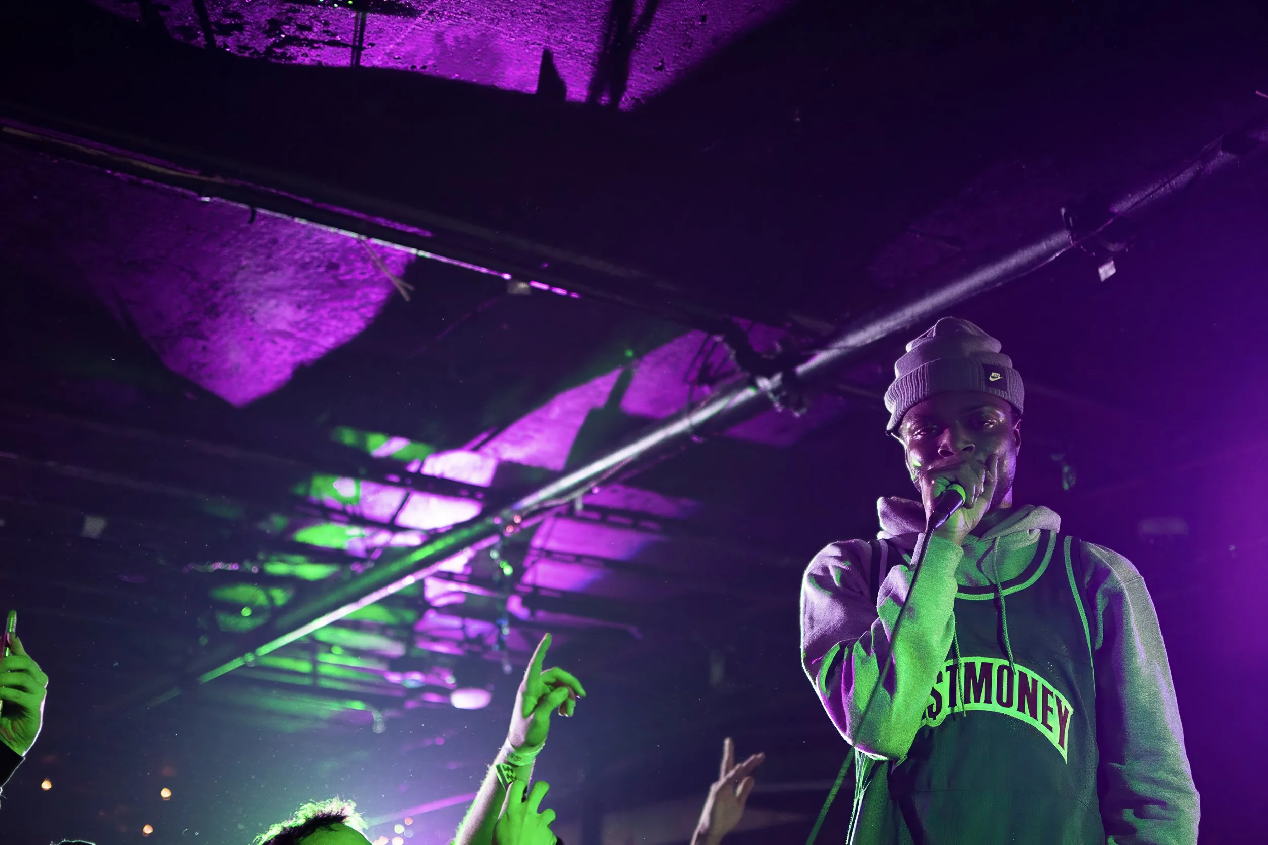 A young man wearing a gray beanie and a neon green hoodie, holding a microphone and performing on stage under purple and green lighting. There are audience members' hands visible in the foreground, some with raised fingers or pointing.