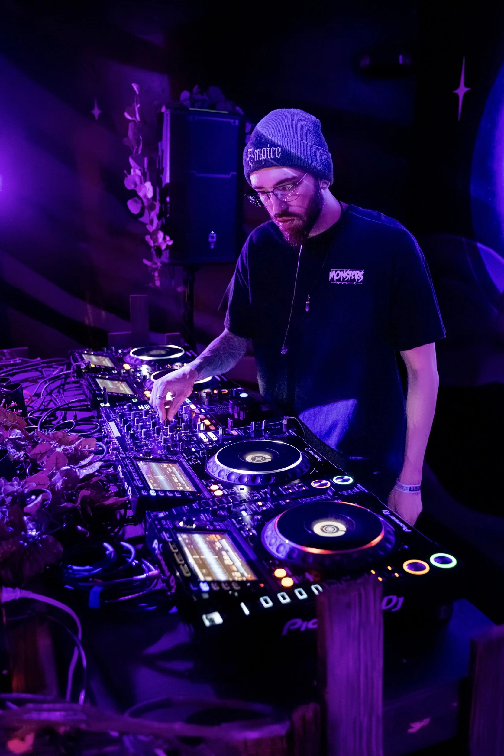 DJ in black T-shirt with 'Monsters' logo, wearing glasses and a gray beanie with 'Empire' text, mixing music on DJ equipment under purple and blue lighting.