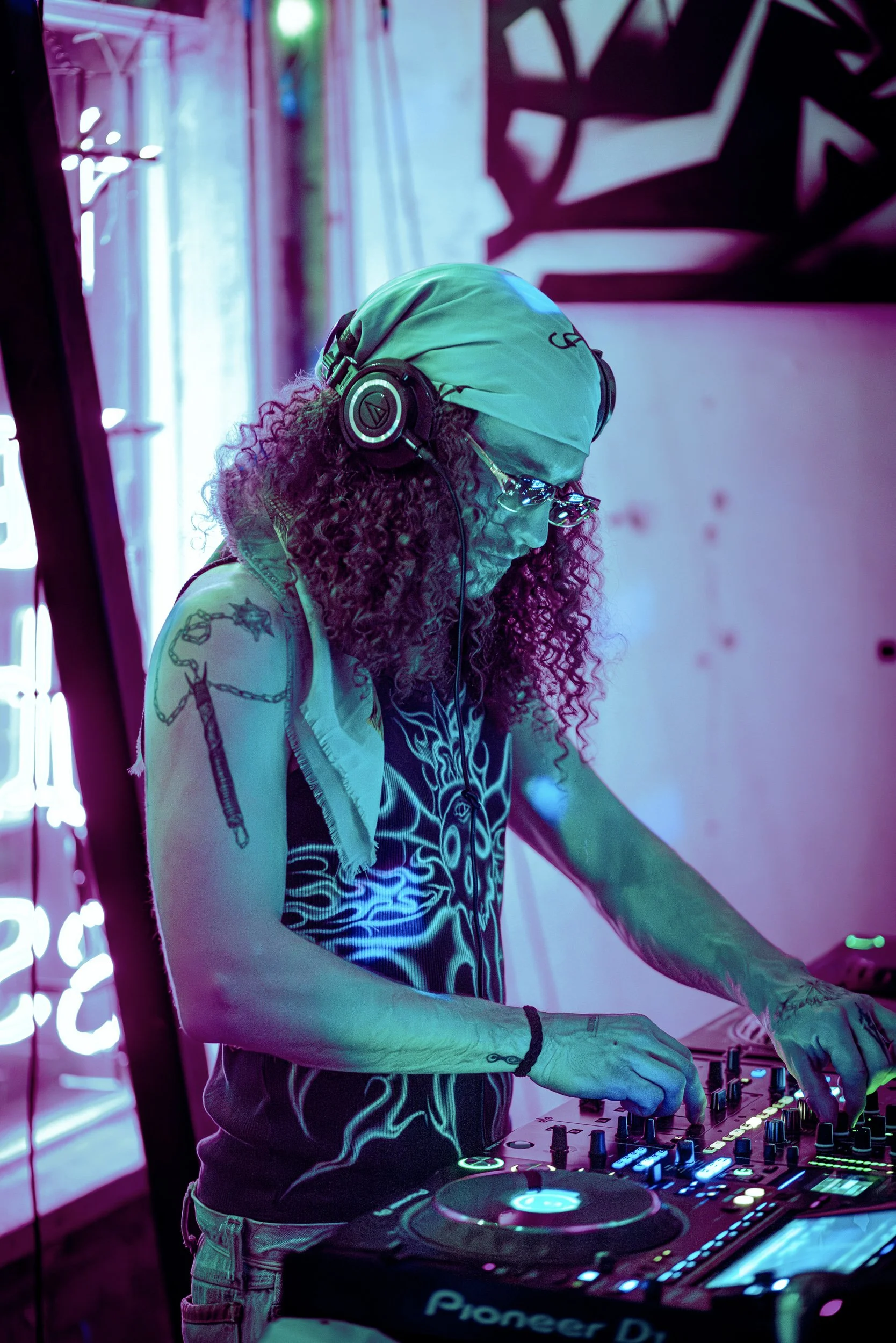 Person DJing with headphones at a neon-lit event, wearing a bandana and sunglasses, operating a Pioneer DJ mixer.