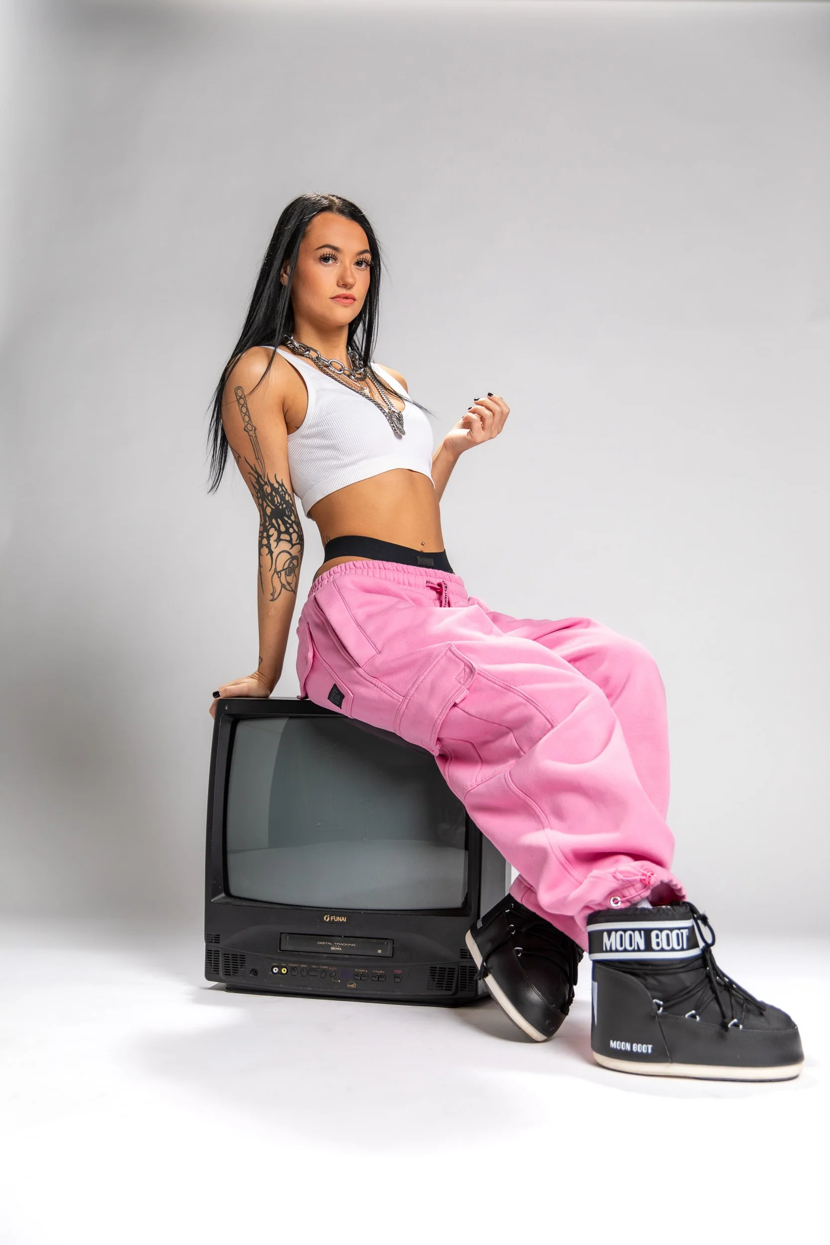 A young woman with long dark hair and tattoos on her arm sitting on an old television set, wearing a white crop top, pink baggy pants, and black moon boot sneakers, against a plain white background.