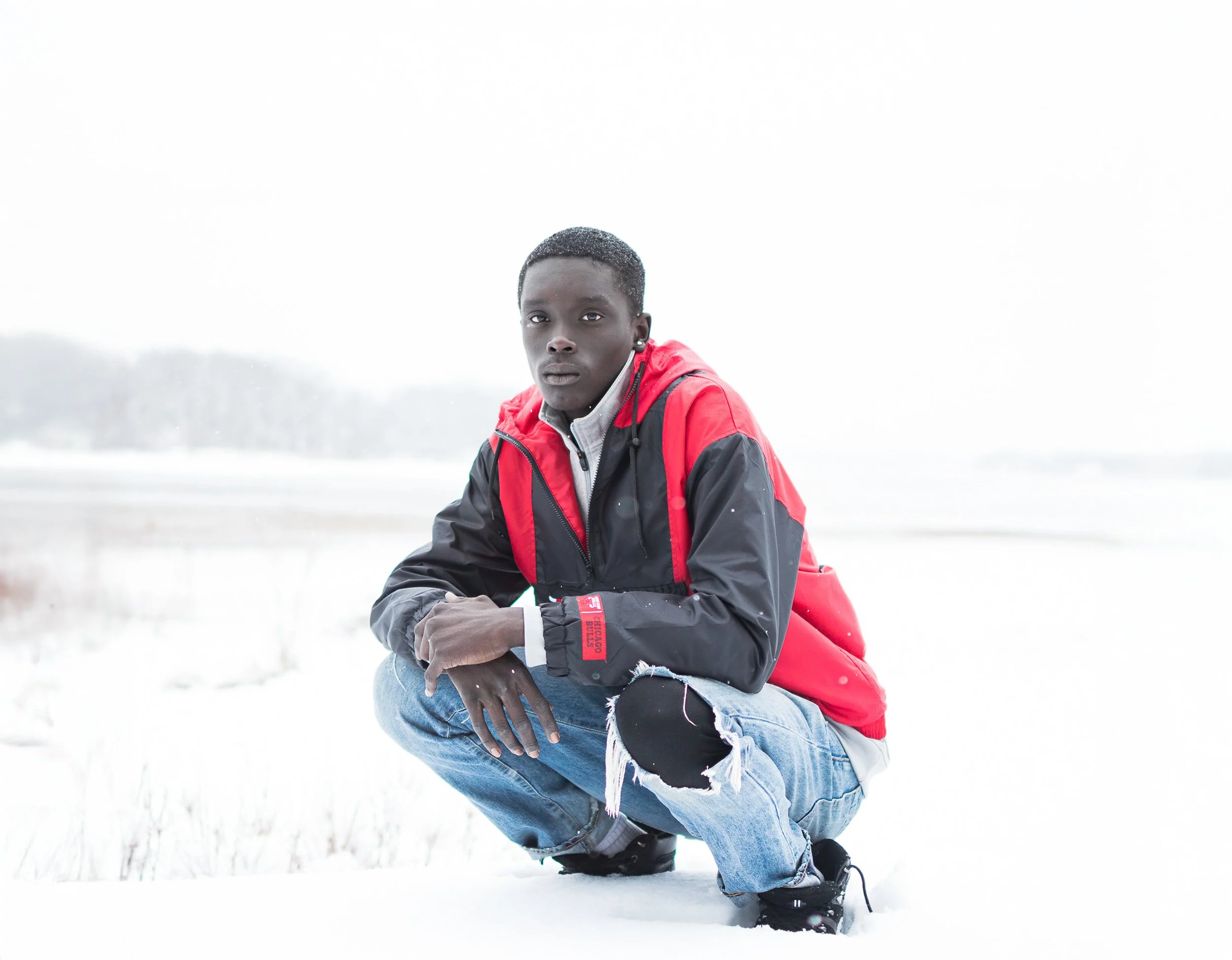 A young man with dark skin and short hair crouches in a snowy landscape, wearing a red and black jacket, distressed jeans, and black boots.