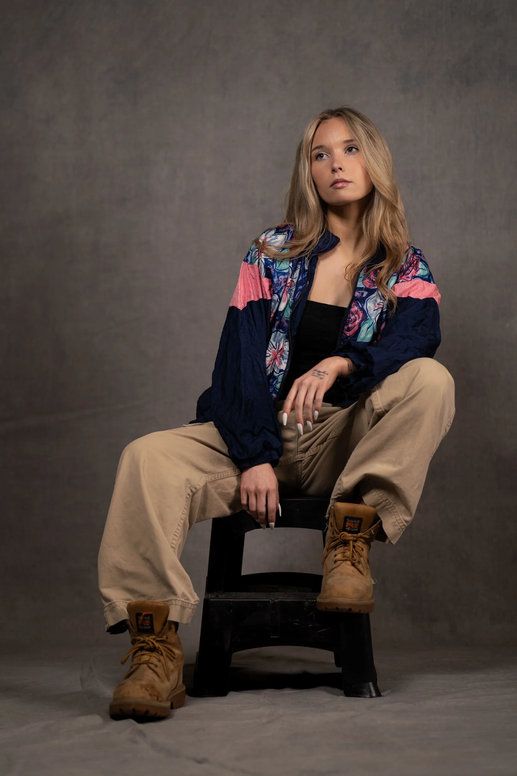 A young woman with long blonde hair sitting on a black chair against a gray background. She wears a colorful floral jacket, black top, beige cargo pants, and tan work boots. She gazes to her left with a neutral expression.