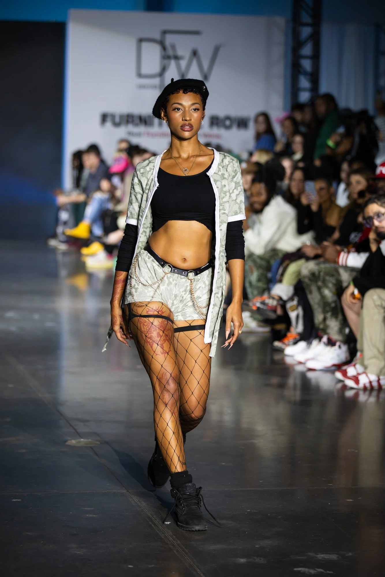 A fashion runway show featuring a female model walking in front of an audience. She is wearing a black crop top, patterned shorts with chain details, fishnet stockings, black combat boots, and a black beret. The background includes a white backdrop w