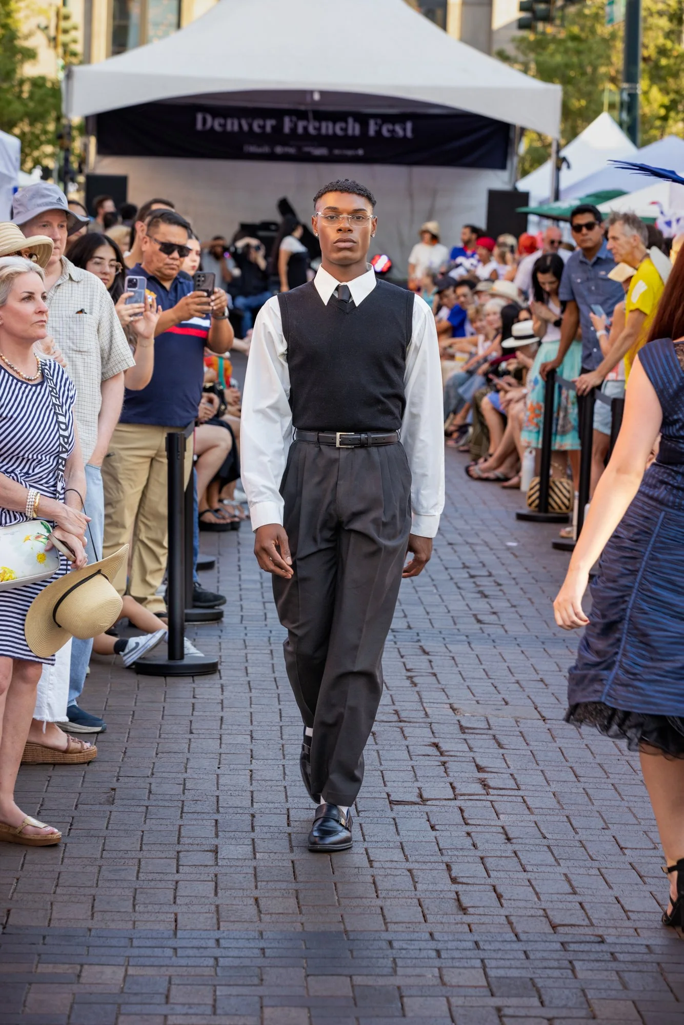 A person walking down a street catwalk at Denver French Fest, surrounded by seated and standing spectators, with a stage and trees in the background.