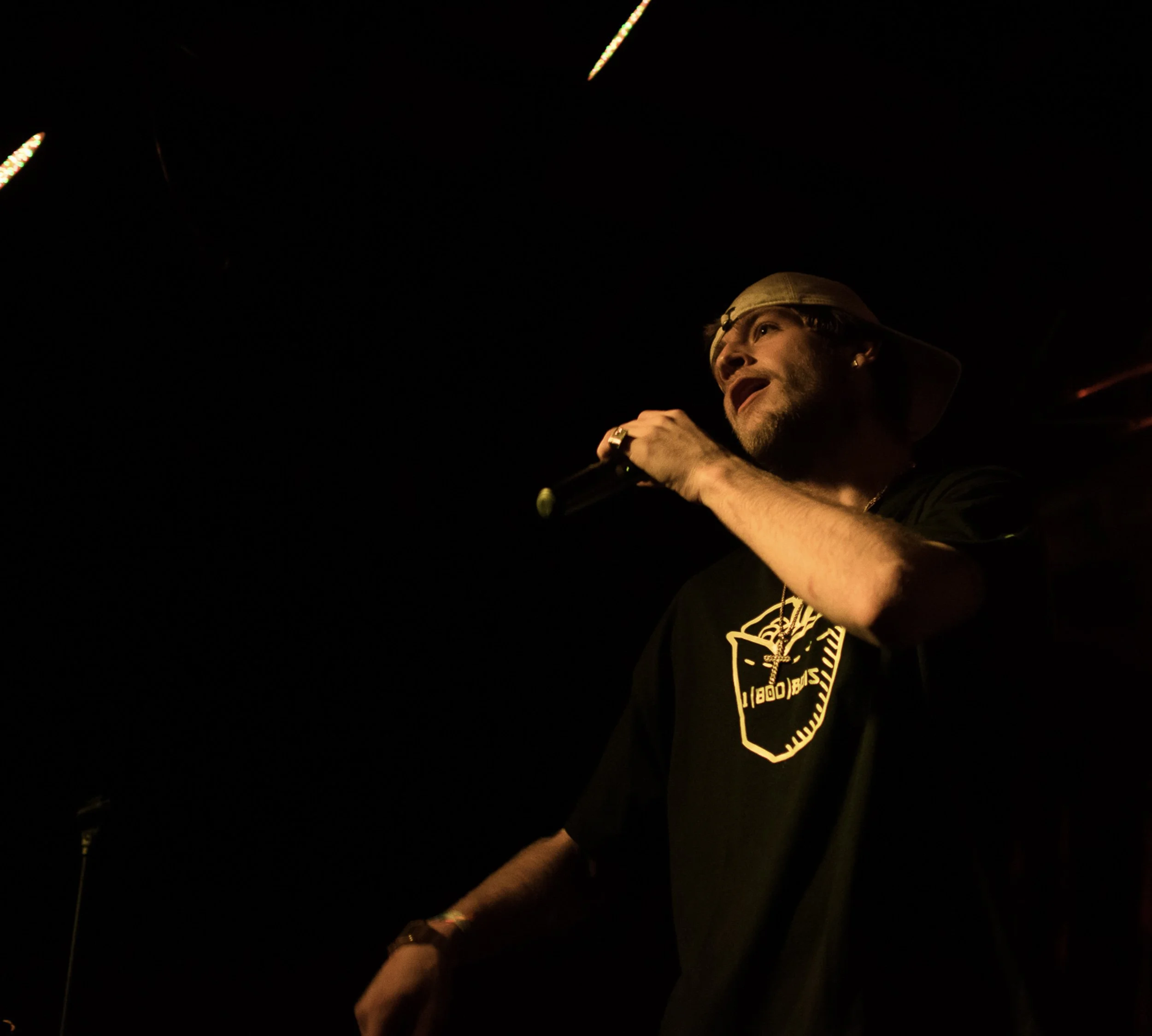 A person wearing a cap and a black shirt with a shield logo is holding a microphone in a dark setting, possibly performing or speaking.