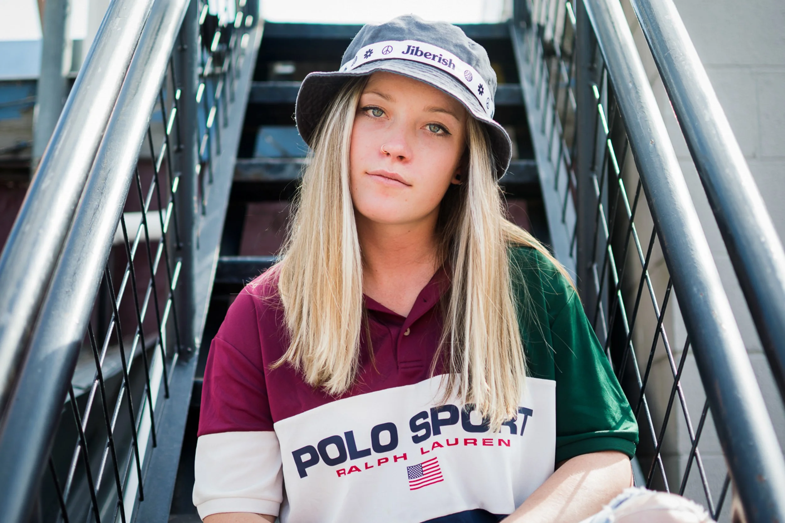 A young woman with long blonde hair wearing a bucket hat and a Polo Sport Ralph Lauren jacket, sitting on outdoor metal stairs.