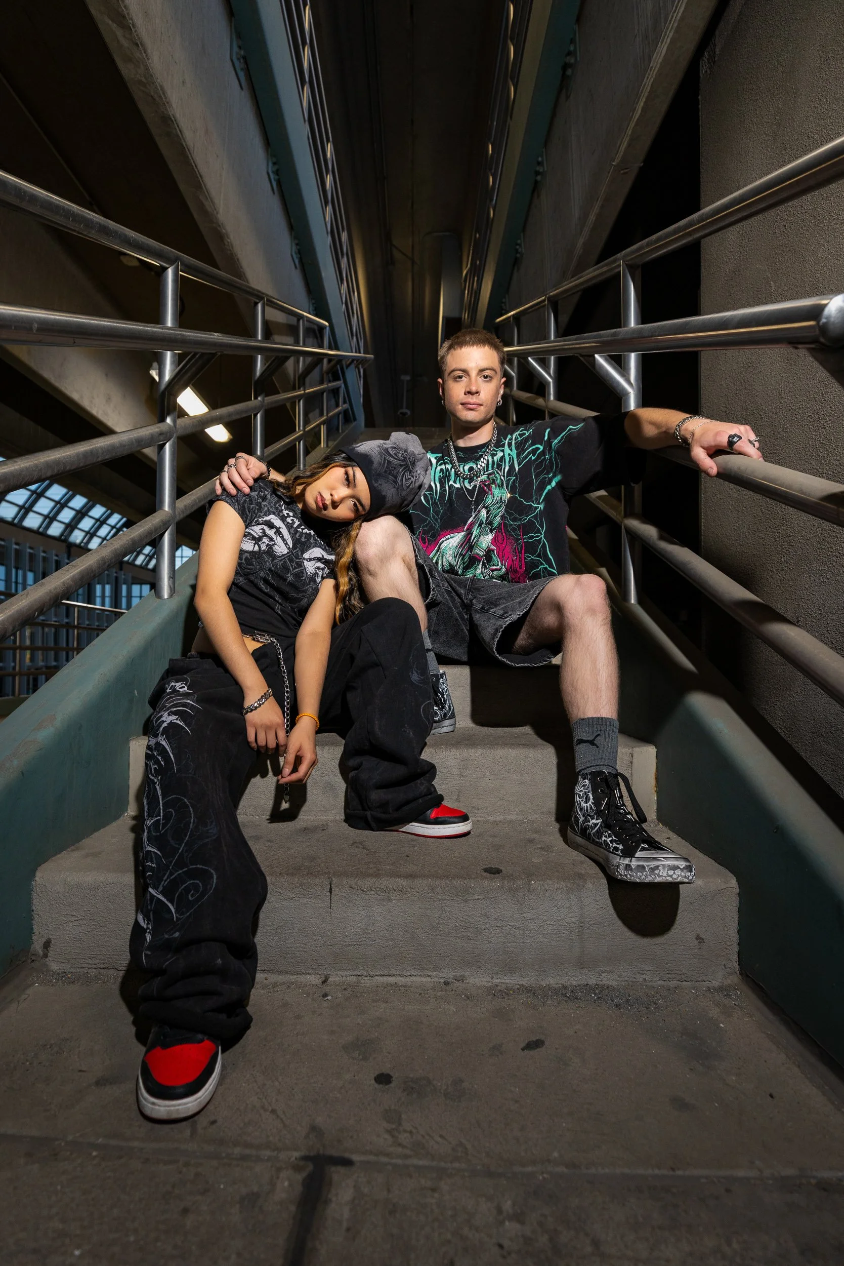 A young woman with long hair, wearing a dark t-shirt and baggy pocketed pants, leaning on a concrete staircase railing with her eyes closed, next to a young man with short hair, wearing a graphic t-shirt and shorts, sitting on the stairs holding both