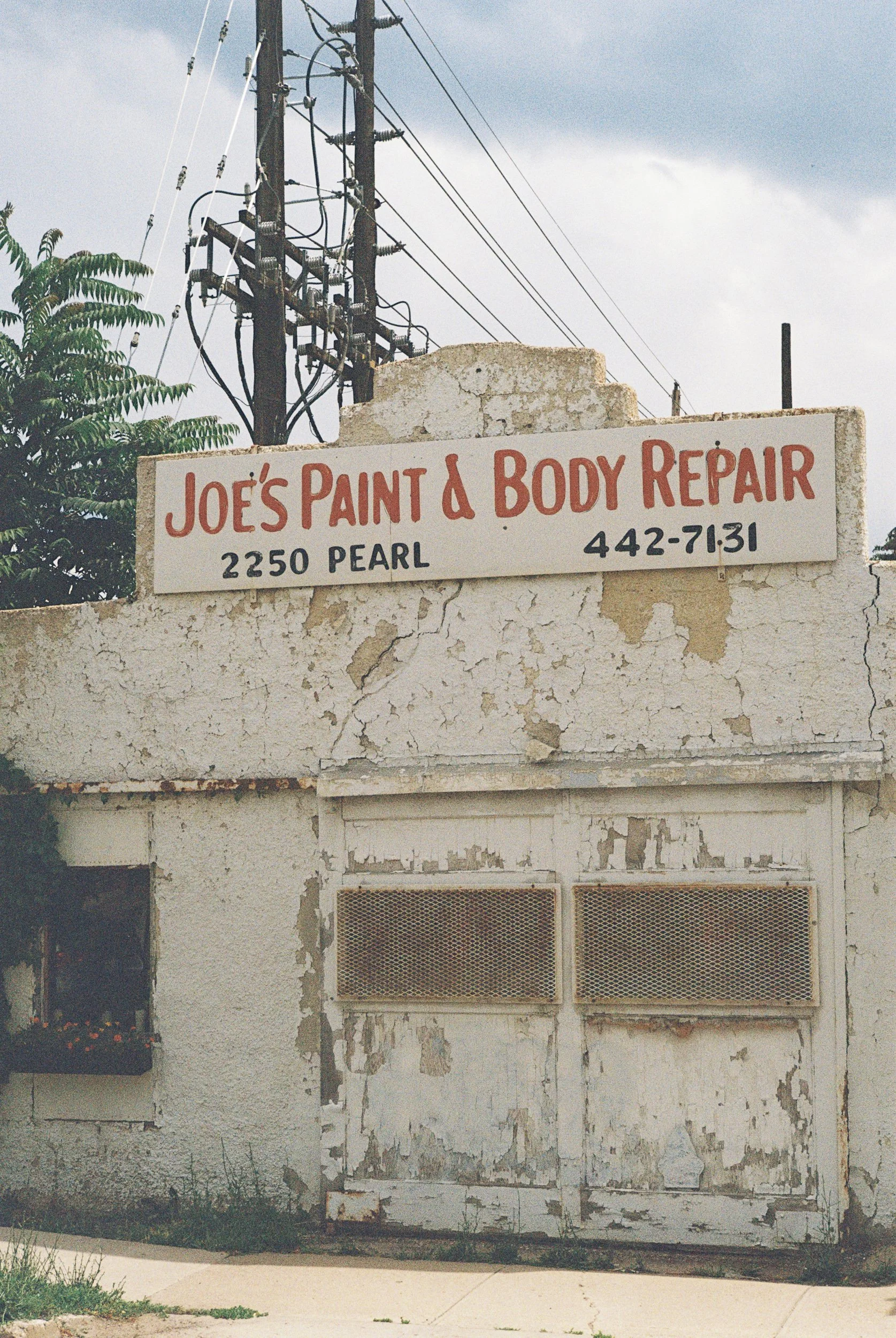 A weathered white sign on a cracked wall reads 'Joe's Paint & Body Repair' with the address '2250 Pearl' and phone number '442-71-31'. Behind the wall are utility poles and wires under a cloudy sky.