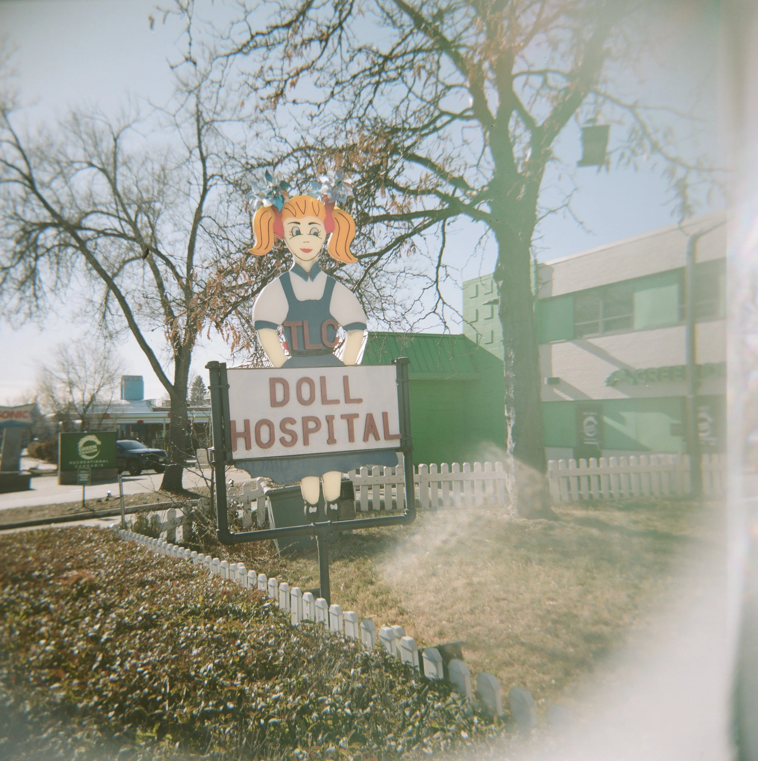 Sign for a doll hospital in front of leafless trees and a building with a green roof during daytime.