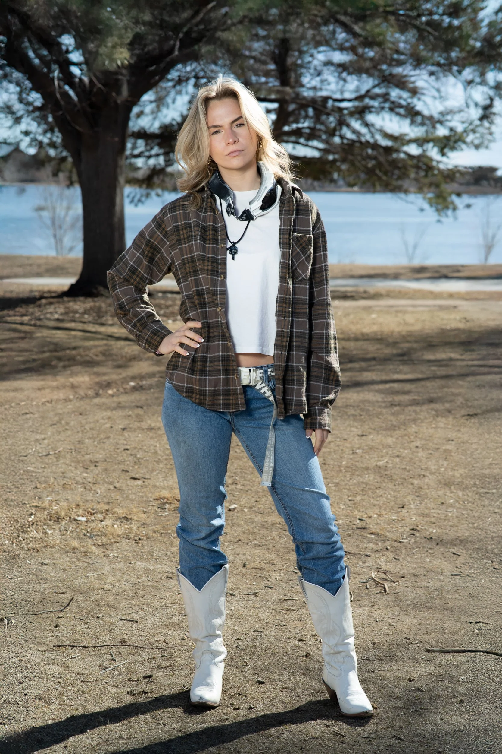 Young woman standing outdoors near a large tree by a body of water, wearing a plaid shirt, white crop top, jeans, white cowboy boots, and a neck scarf with a cross charm.