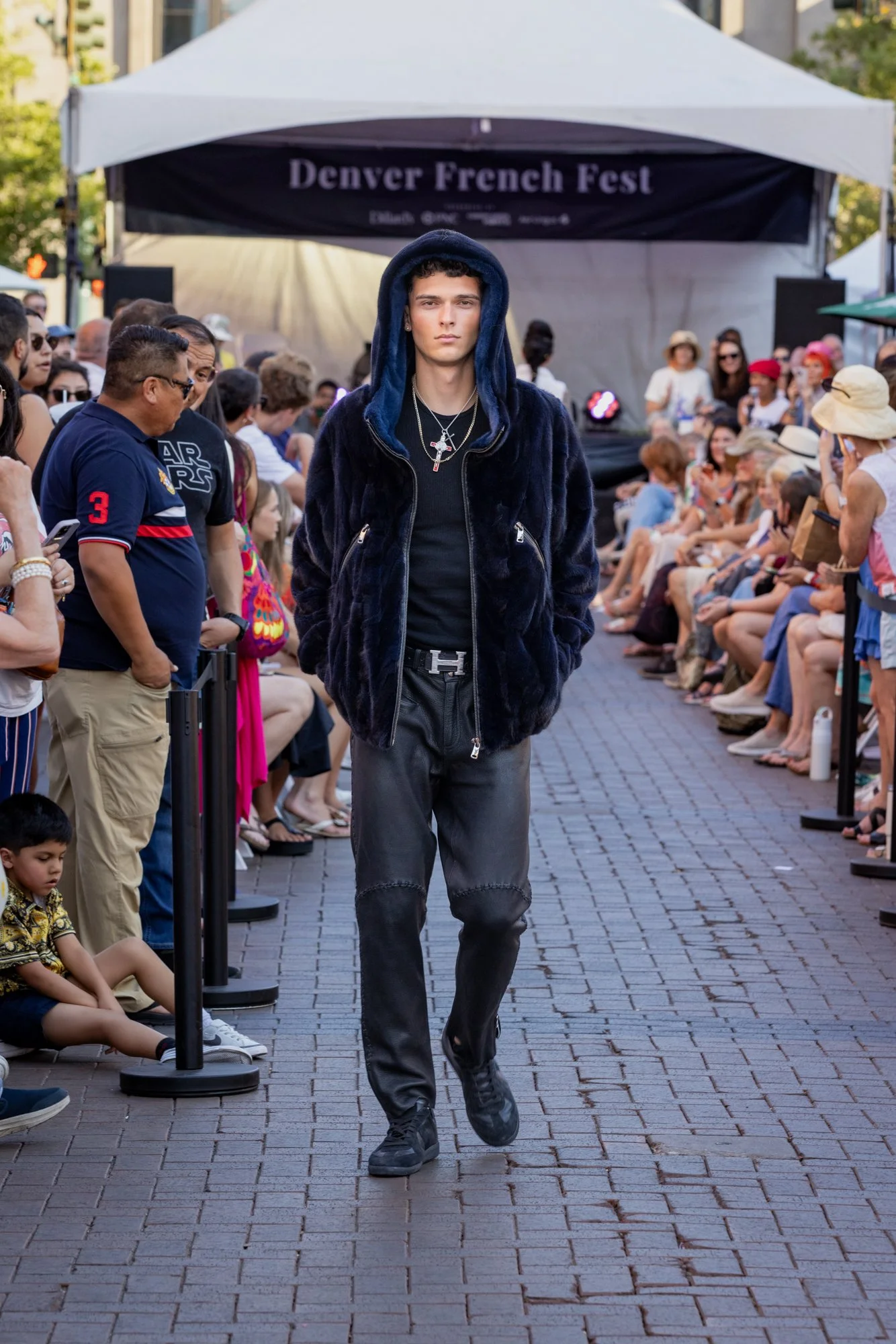 A young male model walks down a cobblestone runway at the Denver French Fest, dressed in dark, casual streetwear including a hooded jacket, baggy jeans, and sneakers, with a crowd of seated spectators watching on both sides.