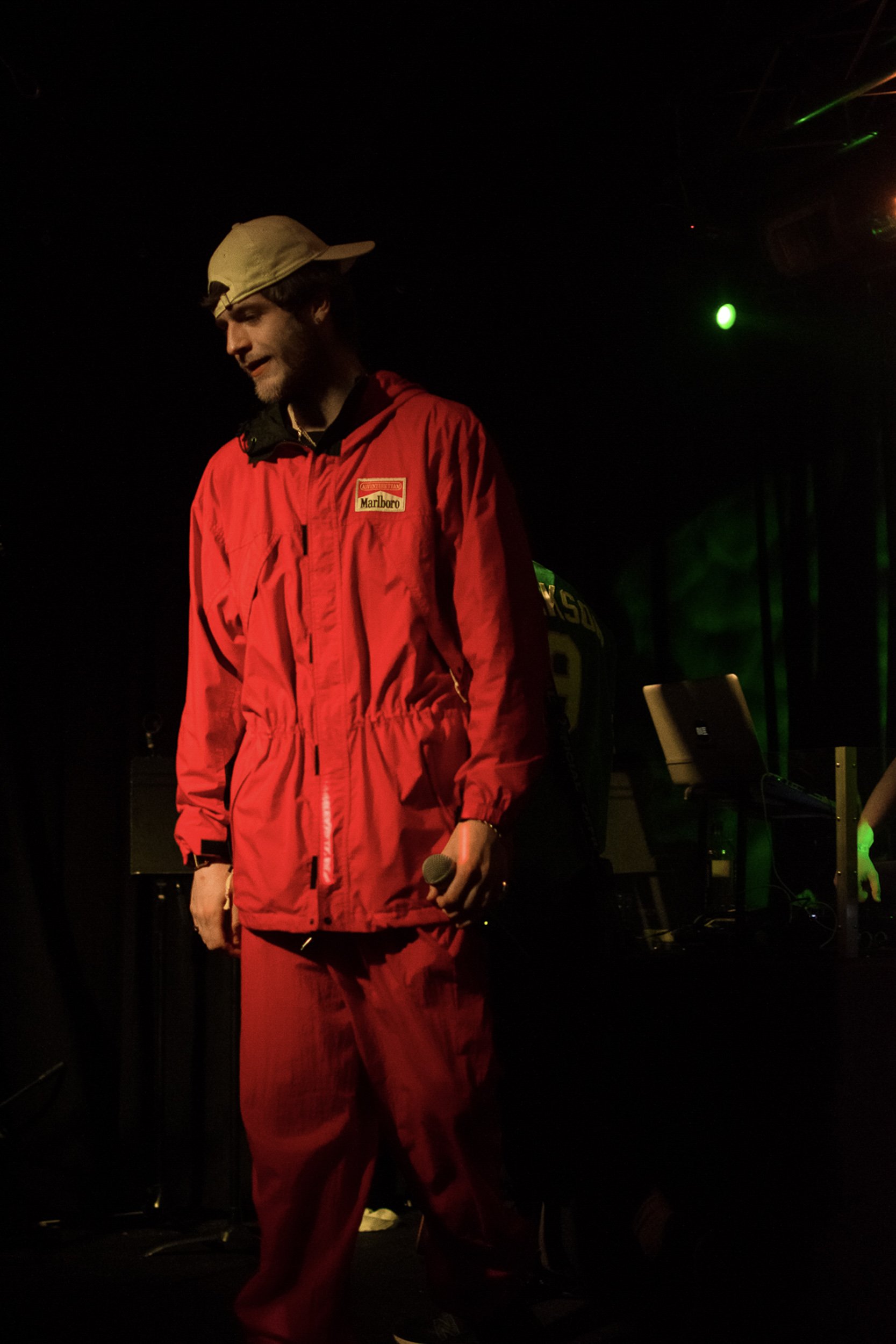 Man in a red jacket and red pants holding a microphone on a dark stage with green and yellow lights.